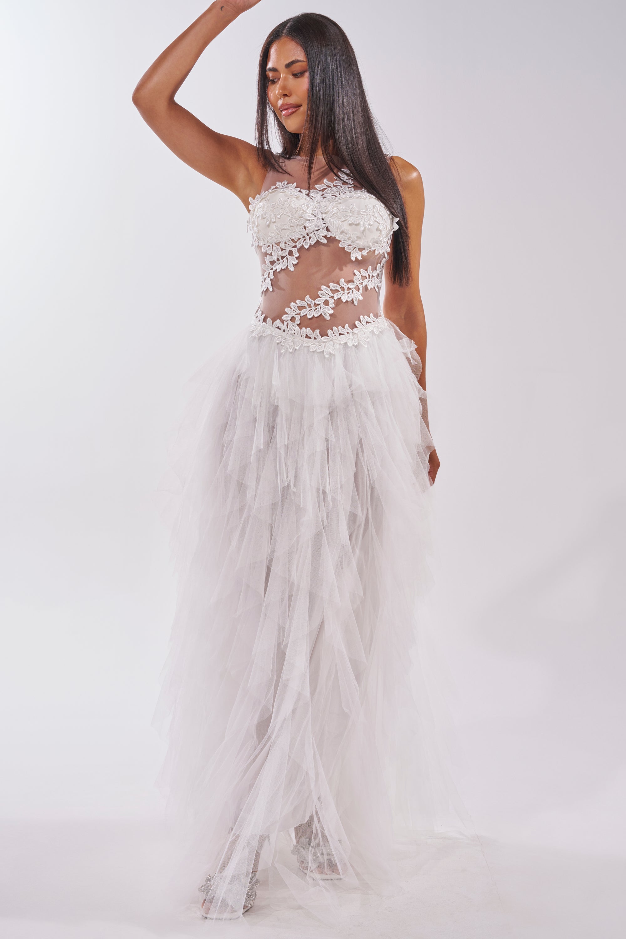A woman poses in the FELT CUTE RUFFLE MAXI DRESS WITH FLORAL APPLIQUES, a sleeveless white dress with a fitted lace bodice, sheer mesh layered tulle skirt, see-through sections, leaf details, and floral appliques. She has long straight hair. 2