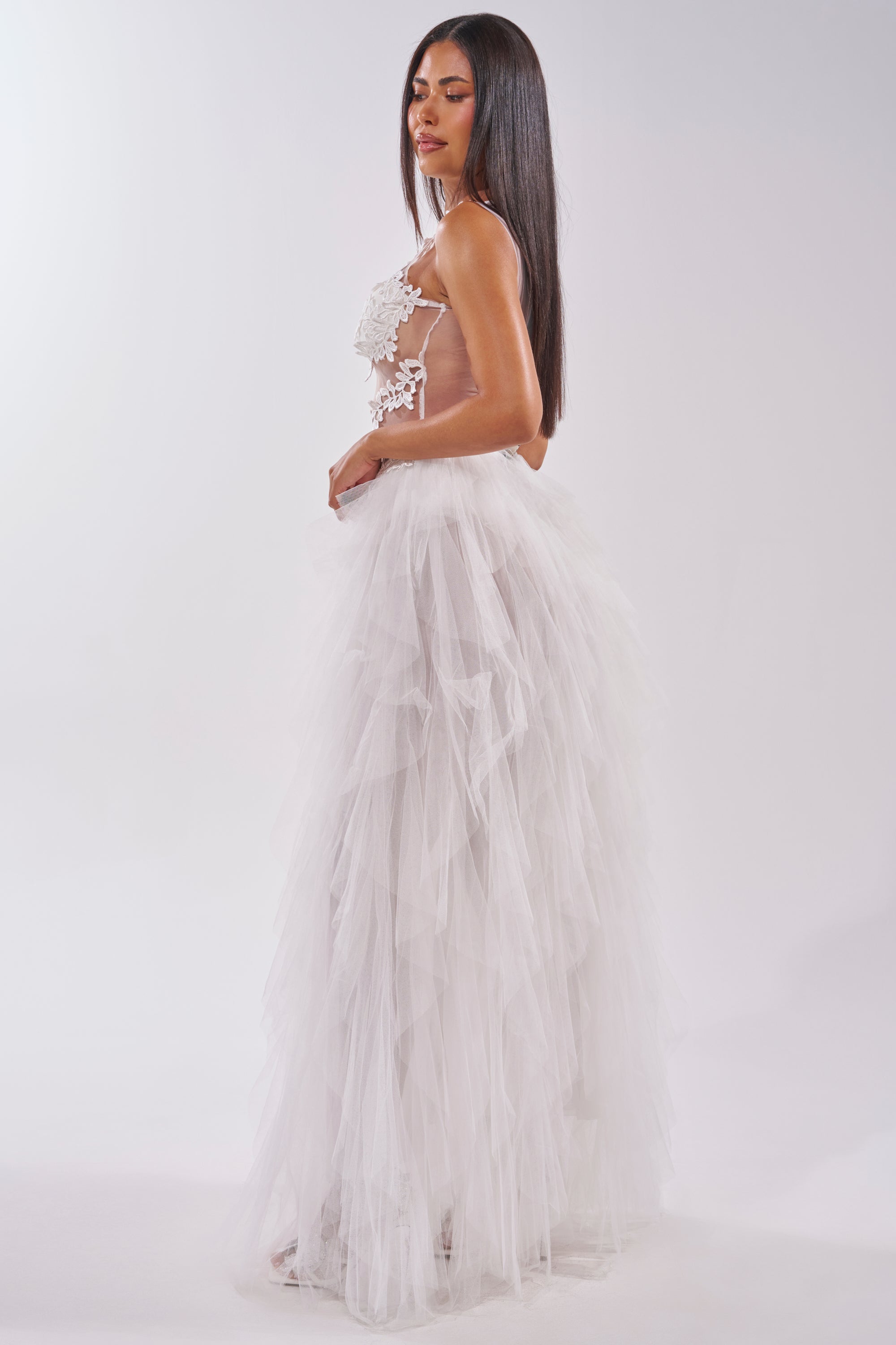 A woman with long straight hair wears the FELT CUTE RUFFLE MAXI DRESS WITH FLORAL APPLIQUES, a sleeveless white gown with a sheer floral-embellished bodice and voluminous tulle skirt, standing sideways against a plain white background. 3