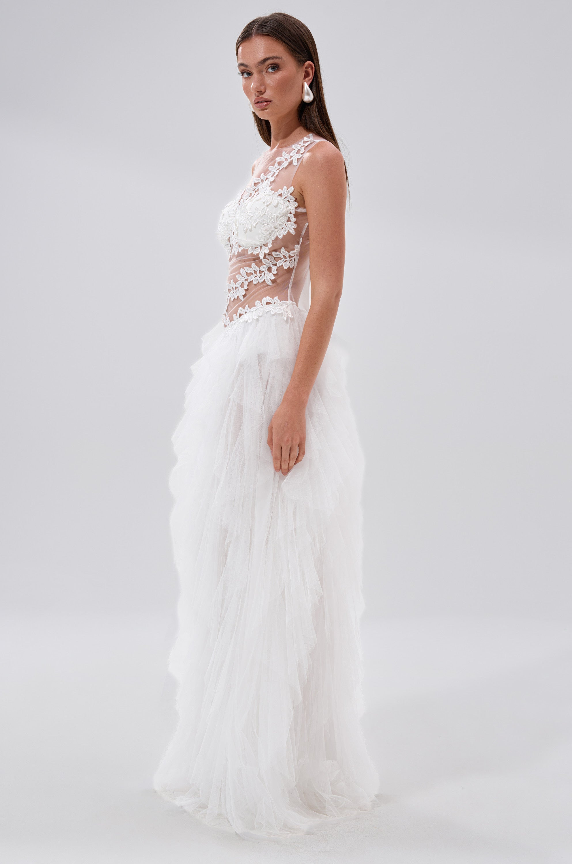 A woman stands sideways in the FELT CUTE RUFFLE MAXI DRESS WITH FLORAL APPLIQUES, featuring a sleeveless white design with a sheer lace bodice and tiered tulle skirt, set against a plain light background.
