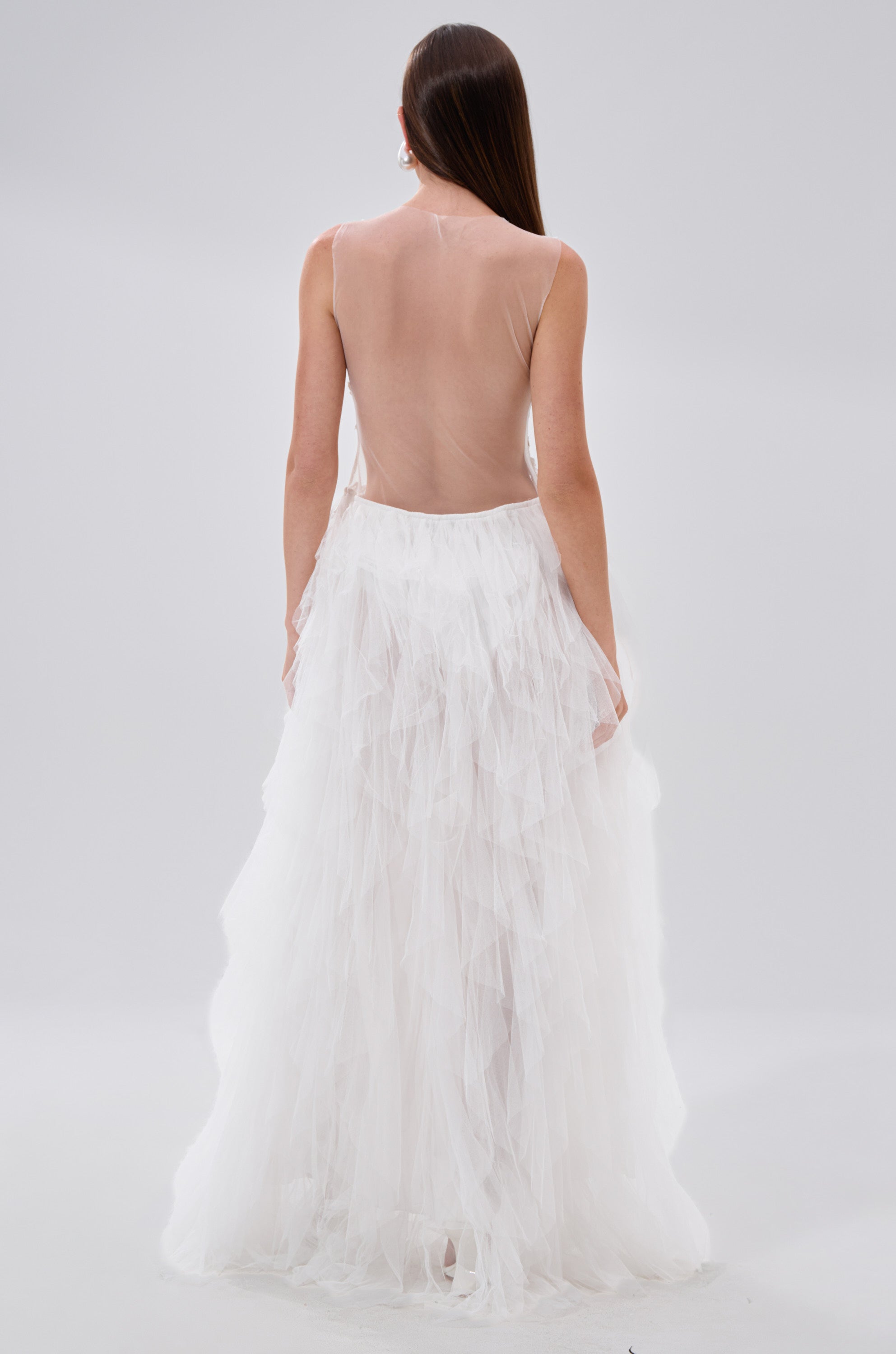A woman faces away, wearing the FELT CUTE RUFFLE MAXI DRESS WITH FLORAL APPLIQUES—a sleeveless, backless white gown with a sheer top and layered tulle skirt. Her straight brown hair flows down her back against a plain light background.