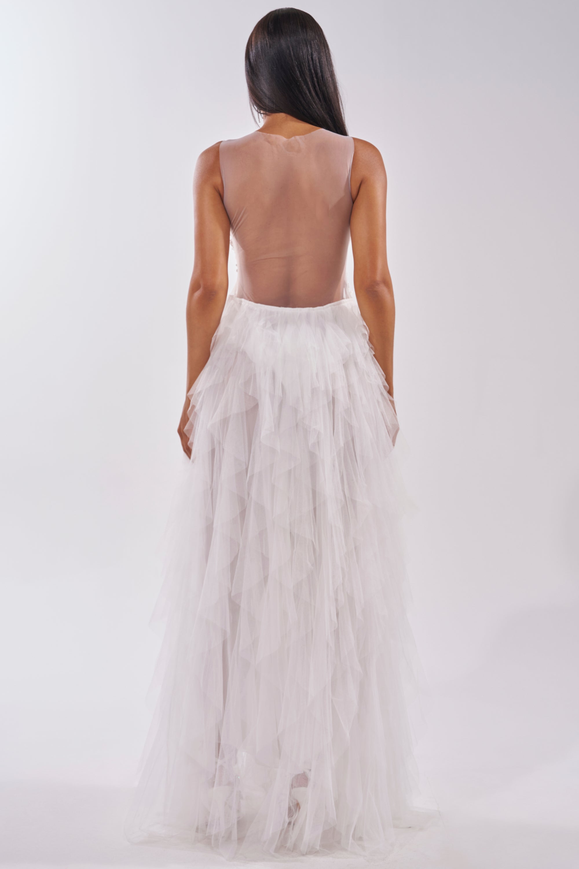 A woman with straight dark hair faces away, wearing the FELT CUTE RUFFLE MAXI DRESS WITH FLORAL APPLIQUES, featuring a sleeveless nude mesh bodice and voluminous layered white tulle skirt, against a plain light background. 5