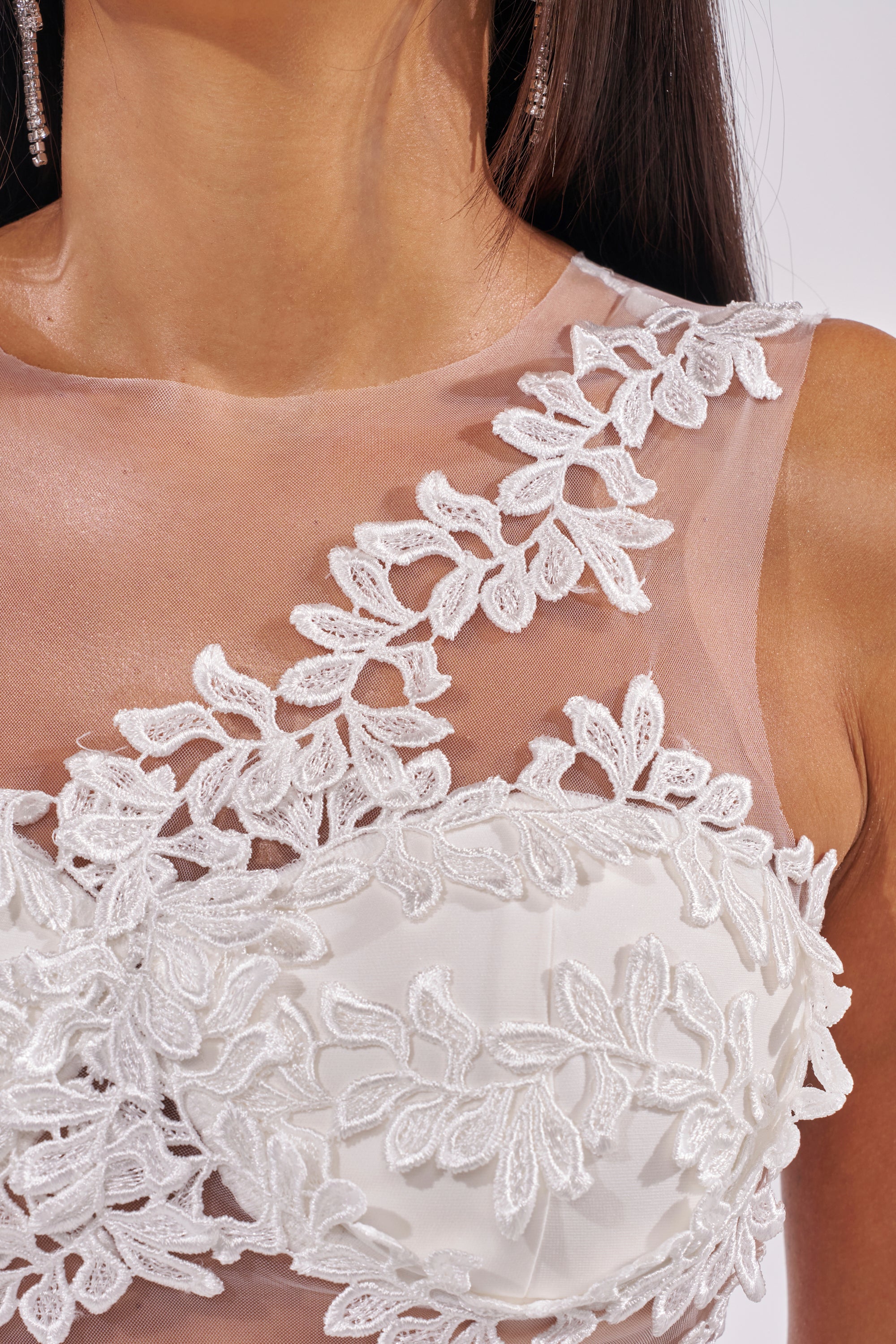 A close-up of a woman in the FELT CUTE RUFFLE MAXI DRESS WITH FLORAL APPLIQUES, featuring a sheer, sleeveless design with white floral lace embroidery and delicate appliques on the bodice and neckline. Her long hair and dangling earrings are partly visible. 7