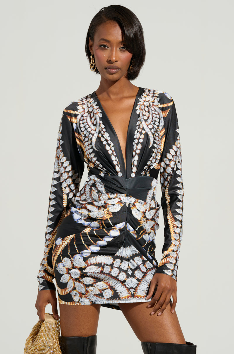 A person wearing the PRINTED ALL OVER MINI DRESS showcases a stylish, long-sleeved look with bold, ornate patterns featuring geometric and feather-like designs. The bodycon silhouette accentuates their figure as they have one hand on their hip and the other holding a gold bag, complemented by knee-high black boots.