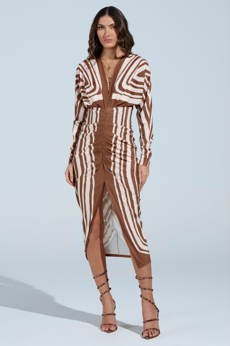 A woman stands against a plain backdrop in the TEXT ME LATER SATIN MIDI DRESS, showcasing its abstract striped pattern and ruched detailing. Her long dark hair flows over the knee-length dress, which she pairs with strappy high-heeled sandals.