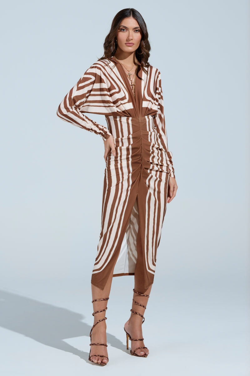 A person confidently wears the "TEXT ME LATER SATIN MIDI DRESS" with brown and white geometric patterns, featuring front ruched detailing, strappy heels, and layered necklaces against a light blue background.