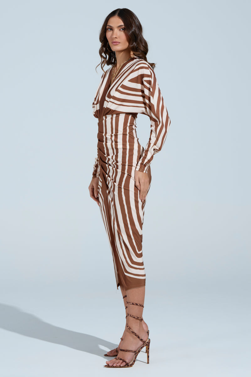 A woman poses in the "TEXT ME LATER SATIN MIDI DRESS," a chic satin piece with an abstract stripe pattern, long sleeves, V-neckline, and mid-calf length. Her strappy high-heel sandals complement her cascading wavy hair against the plain light blue backdrop.