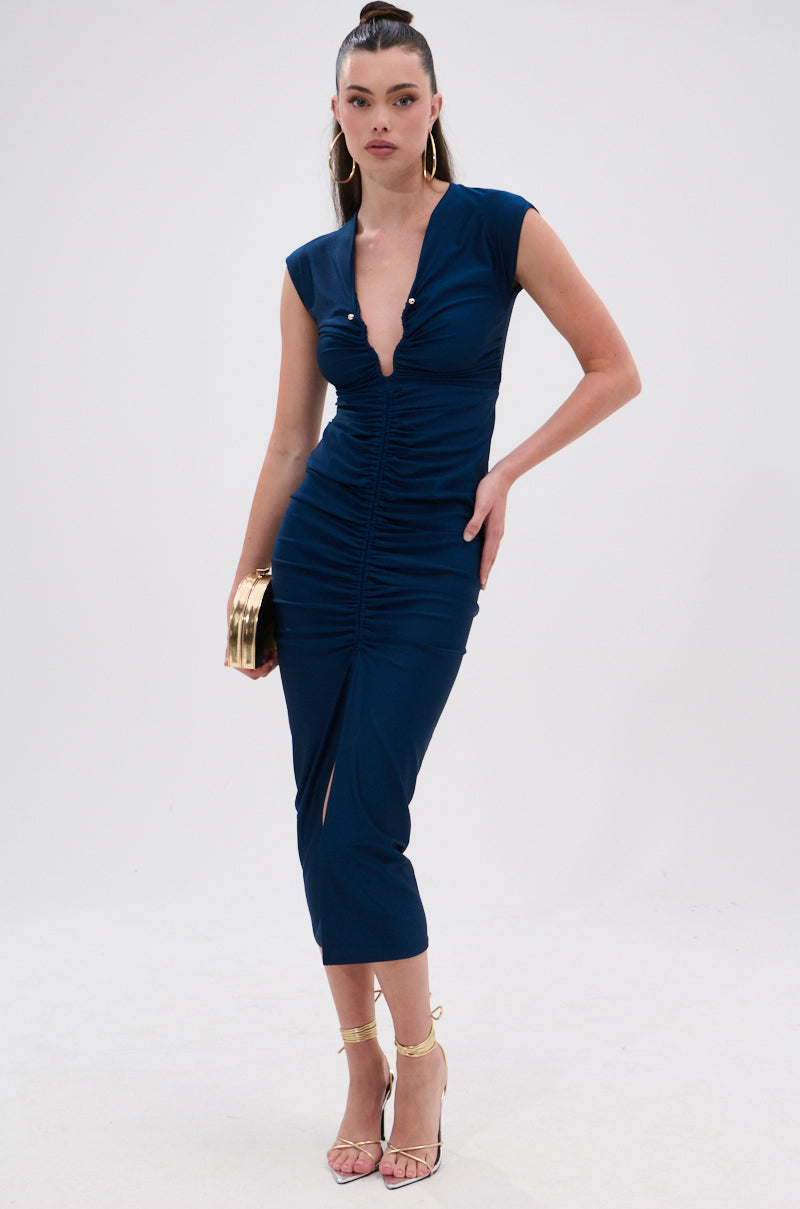A woman wears the JULIE SLINKY MIDI DRESS in deep blue, featuring a curve-hugging fit, cap sleeves, and a plunging neckline. She pairs it with hoop earrings, strappy heels, and a gold clutch against a plain white background. 1