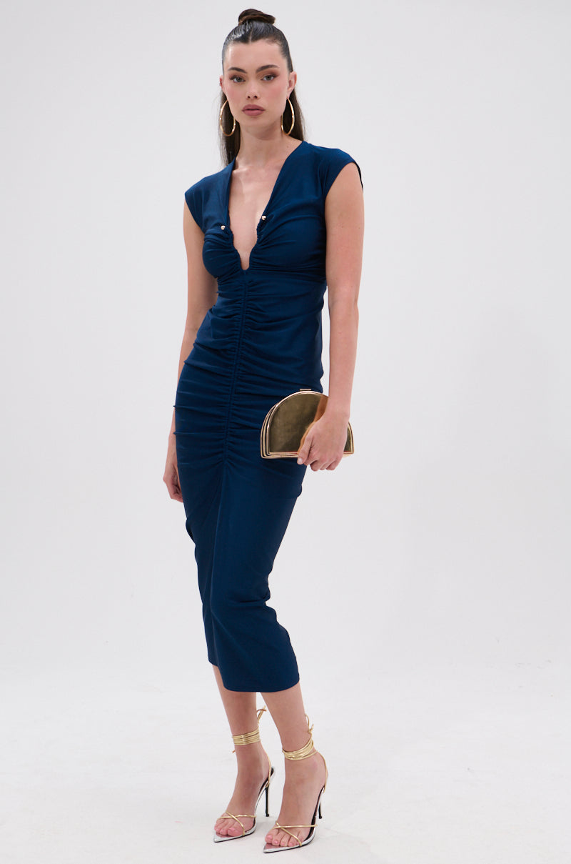 A woman stands against a plain background wearing the JULIE SLINKY MIDI DRESS, styled with gold hoop earrings, a gold bracelet, strappy gold heels, and a small gold clutch. Her hair is pulled back in a sleek ponytail. 2
