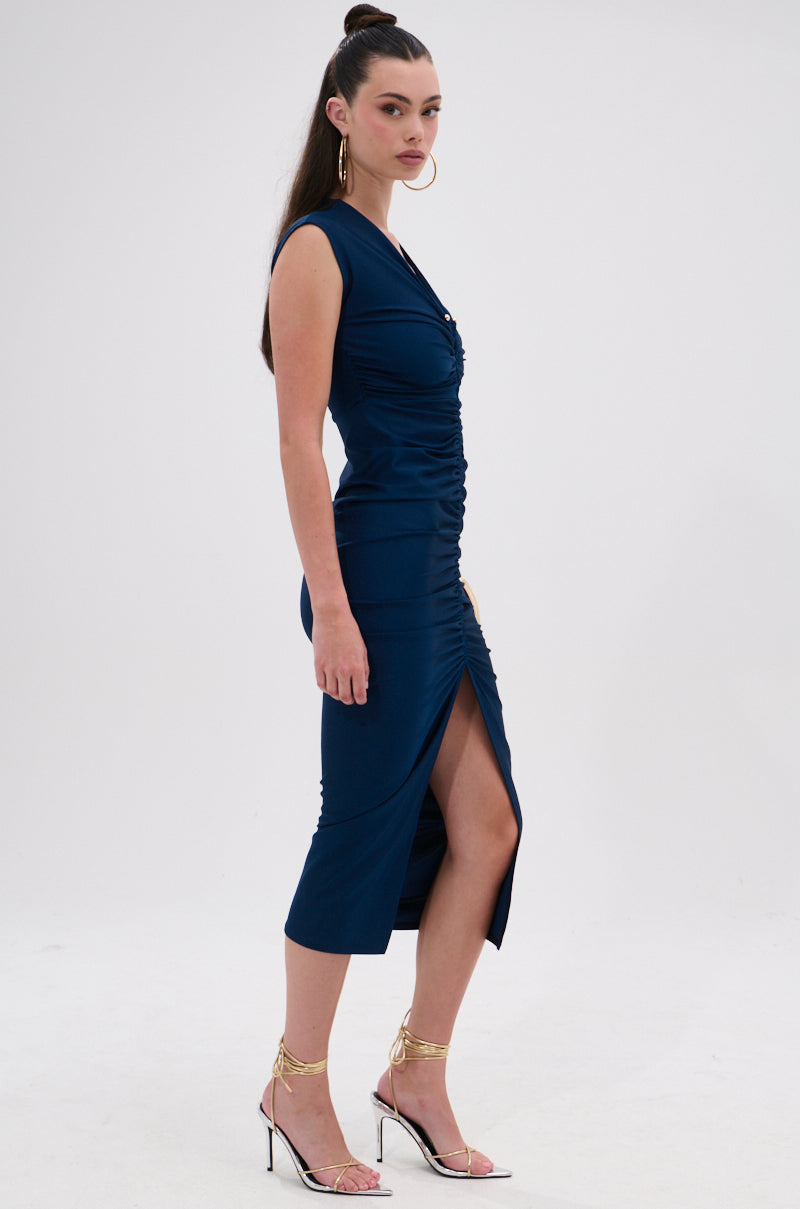 A woman stands in profile wearing the JULIE SLINKY MIDI DRESS with a curve-hugging fit and front slit. She pairs it with clear high-heeled sandals featuring gold straps, and her long dark hair is styled in a high ponytail. 4
