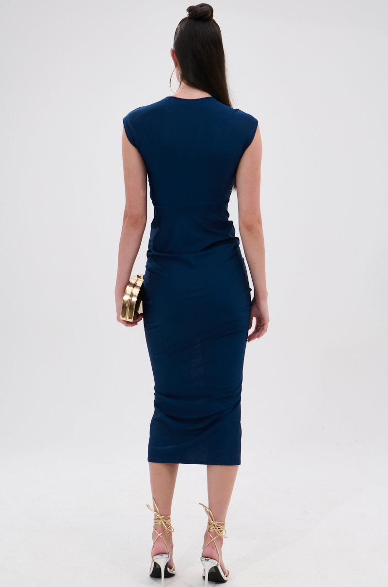 A woman faces away in the curve-hugging JULIE SLINKY MIDI DRESS, a fitted navy blue knee-length style. She holds a gold clutch, wears her hair in a topknot, and sports white high-heeled sandals with gold straps. 5