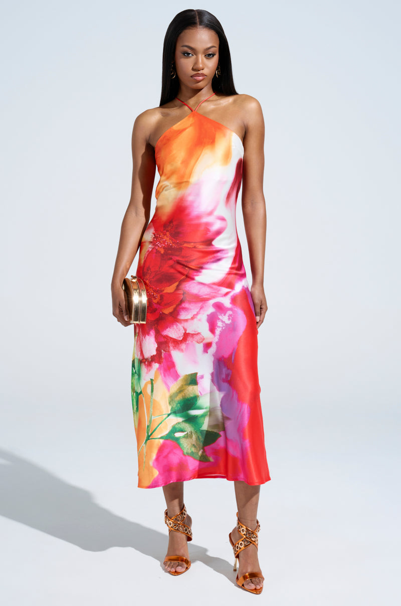 Dressed in the TIME MOVES FAST PRINTED MIDI DRESS, a woman showcases a vibrant, abstract floral print with a halter neckline in red, orange, and pink hues. She holds a small metallic clutch and dons strappy high heels against a plain, light background.