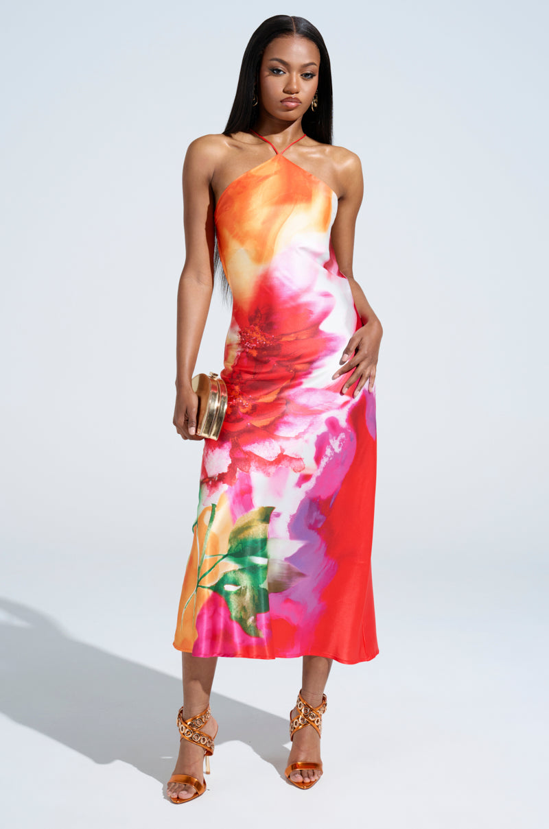 The woman stands elegantly in the TIME MOVES FAST PRINTED MIDI DRESS, featuring a vibrant abstract floral print and open back. She holds a gold clutch against a light gray backdrop, completing her chic look with strappy heels.