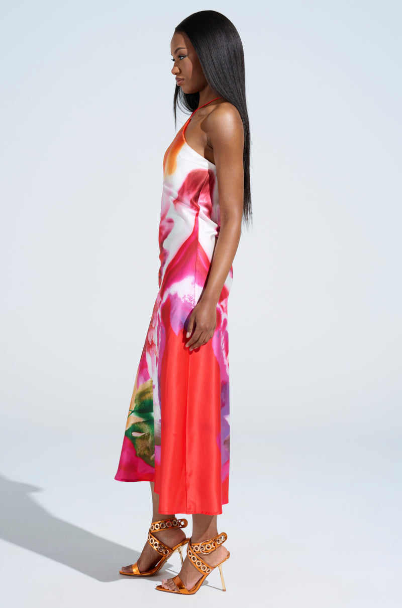 A person with long, straight hair dons the TIME MOVES FAST PRINTED MIDI DRESS, showcasing an abstract floral print and halter neckline. They pair it flawlessly with decorative high-heeled sandals against a plain, light-colored background.
