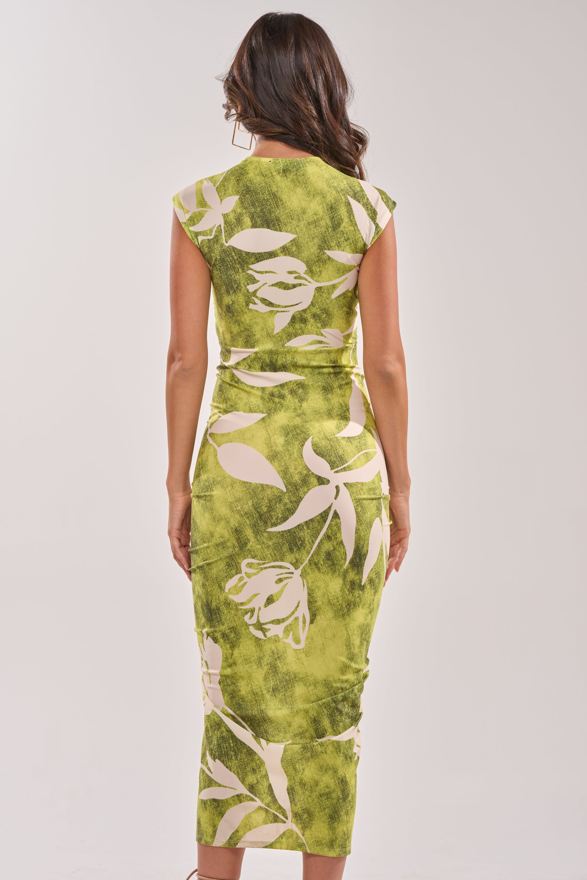 A woman stands with her back to the camera against a plain light background, modeling the FUN IN THE SUN CINCHED MIDI DRESS—a fitted green tropical dress with cap sleeves and a below-the-knee length. 4