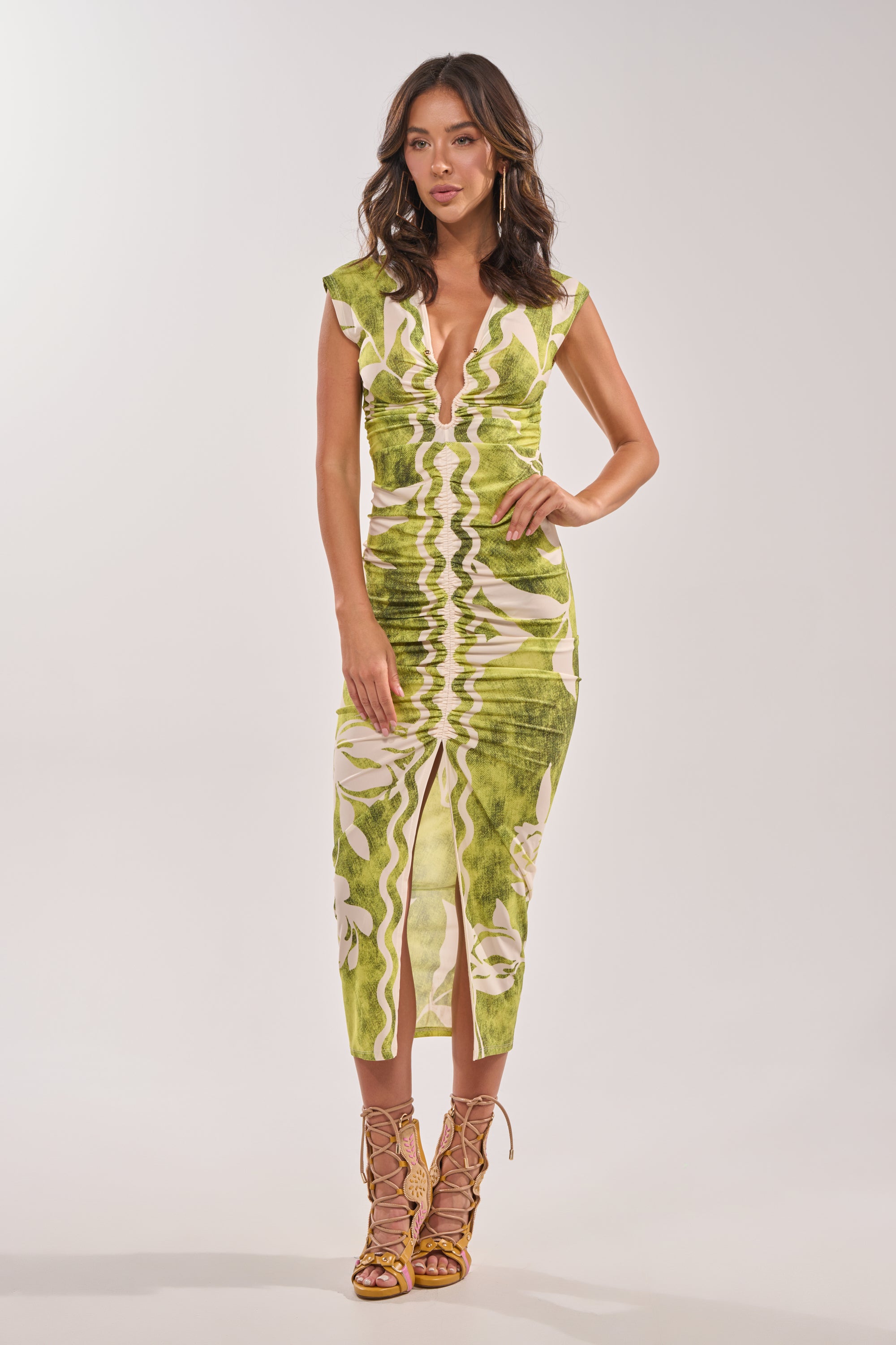 A woman wears the FUN IN THE SUN CINCHED MIDI DRESS, featuring a green tropical print, plunging V-neckline, and ruched details. She pairs it with strappy nude heels against a plain light background. 5
