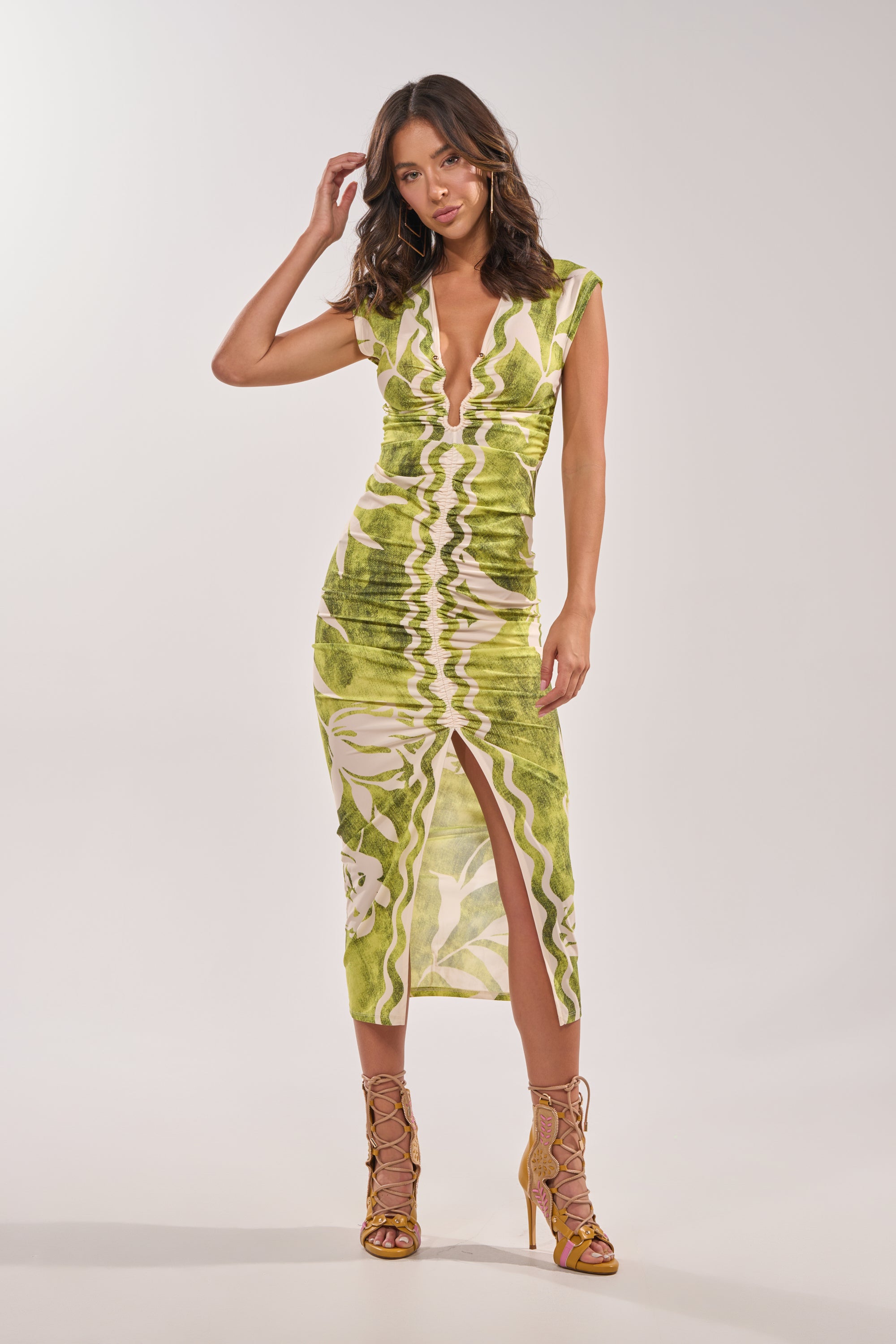 A woman poses against a light backdrop, wearing the FUN IN THE SUN CINCHED MIDI DRESS in green with a ruched bodycon fit, plunging V-neckline, front slit, and strappy yellow high-heeled sandals. 6