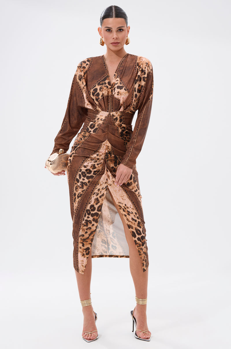 A woman faces forward in the LOTS OF LOVE TO GIVE MIDI DRESS, featuring leopard patchwork, long sleeves, and a front slit. She accessorizes with hoop earrings, slicked-back hair, and gold heeled sandals against a plain white background.