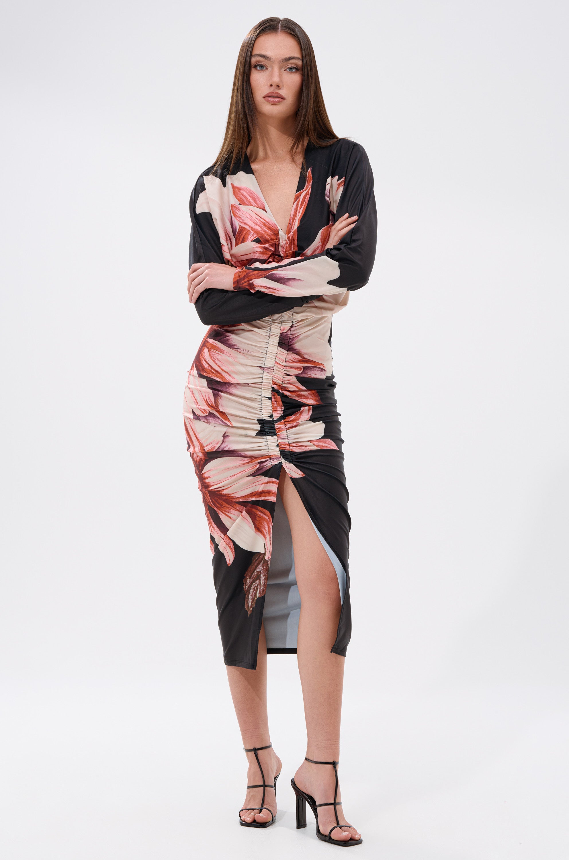 A woman with straight brown hair poses with arms crossed in the FLORAL BABY MIDI DRESS, which features a floral print, bodycon blouson silhouette, and front slit. She completes her look with black high-heeled sandals. 0