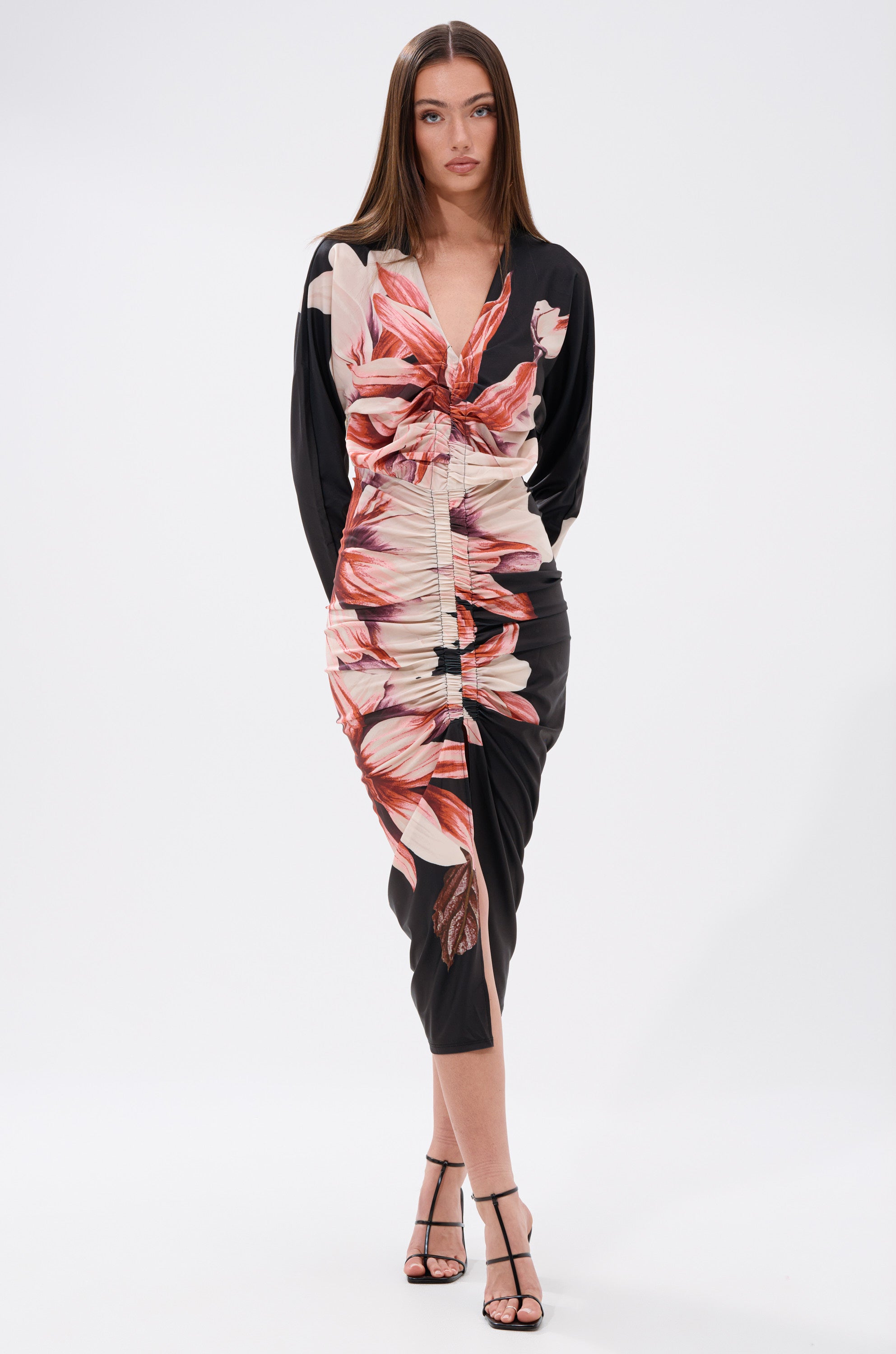 A woman stands against a plain background wearing the FLORAL BABY MIDI DRESS, a black long-sleeve ruched bodycon featuring bold red and beige floral prints, paired with strappy black heels. She faces forward with arms relaxed at her sides. 1