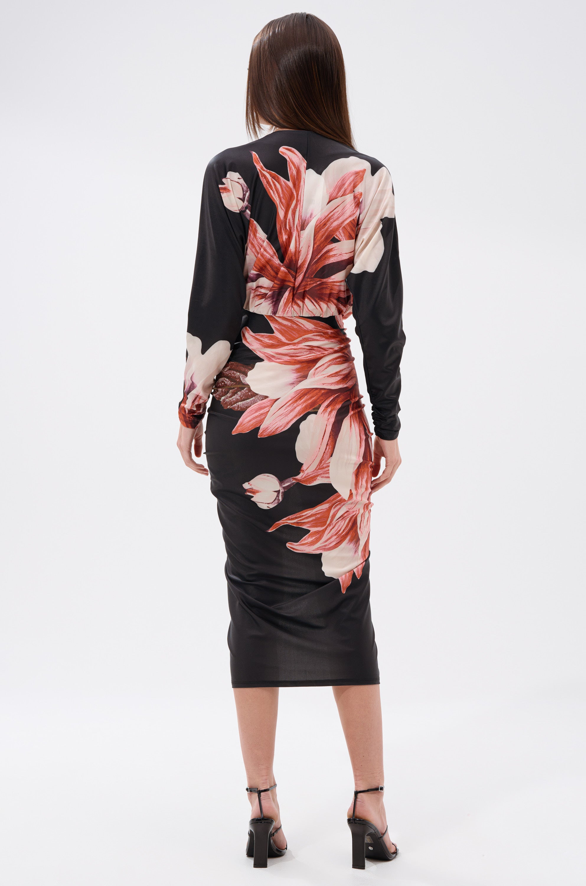 A woman with straight brown hair faces away, modeling the FLORAL BABY MIDI DRESS—a body-hugging floral piece with large pink and white patterns—paired with black heeled sandals against a plain white background. 3