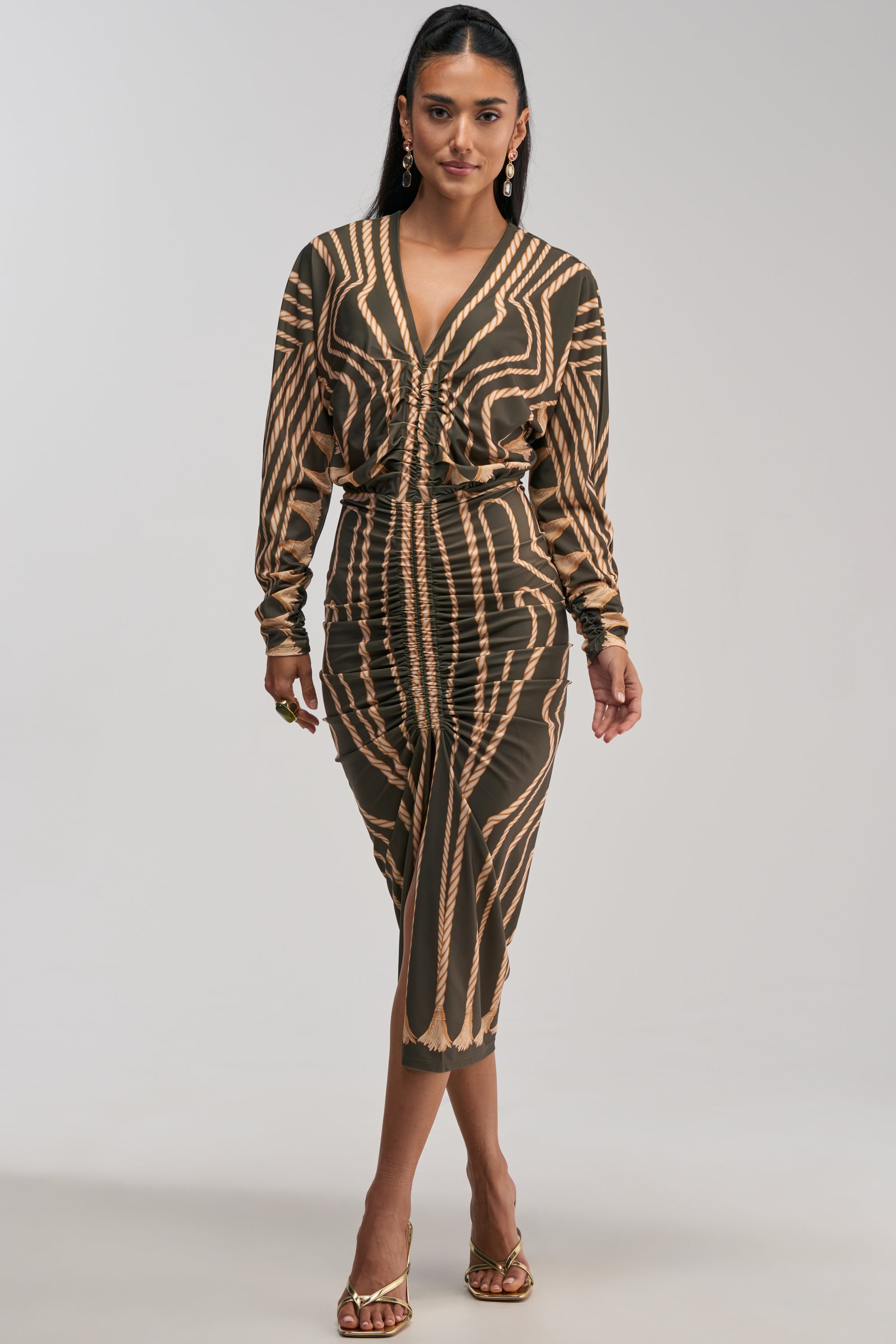 A woman wearing the RESERVATIONS ONLY PRINTED MIDI DRESS IN OLIVE, featuring long sleeves and a knee-length abstract print with tan and dark brown geometric patterns, styled with gold earrings, rings, and open-toe heels, stands against a plain light background.