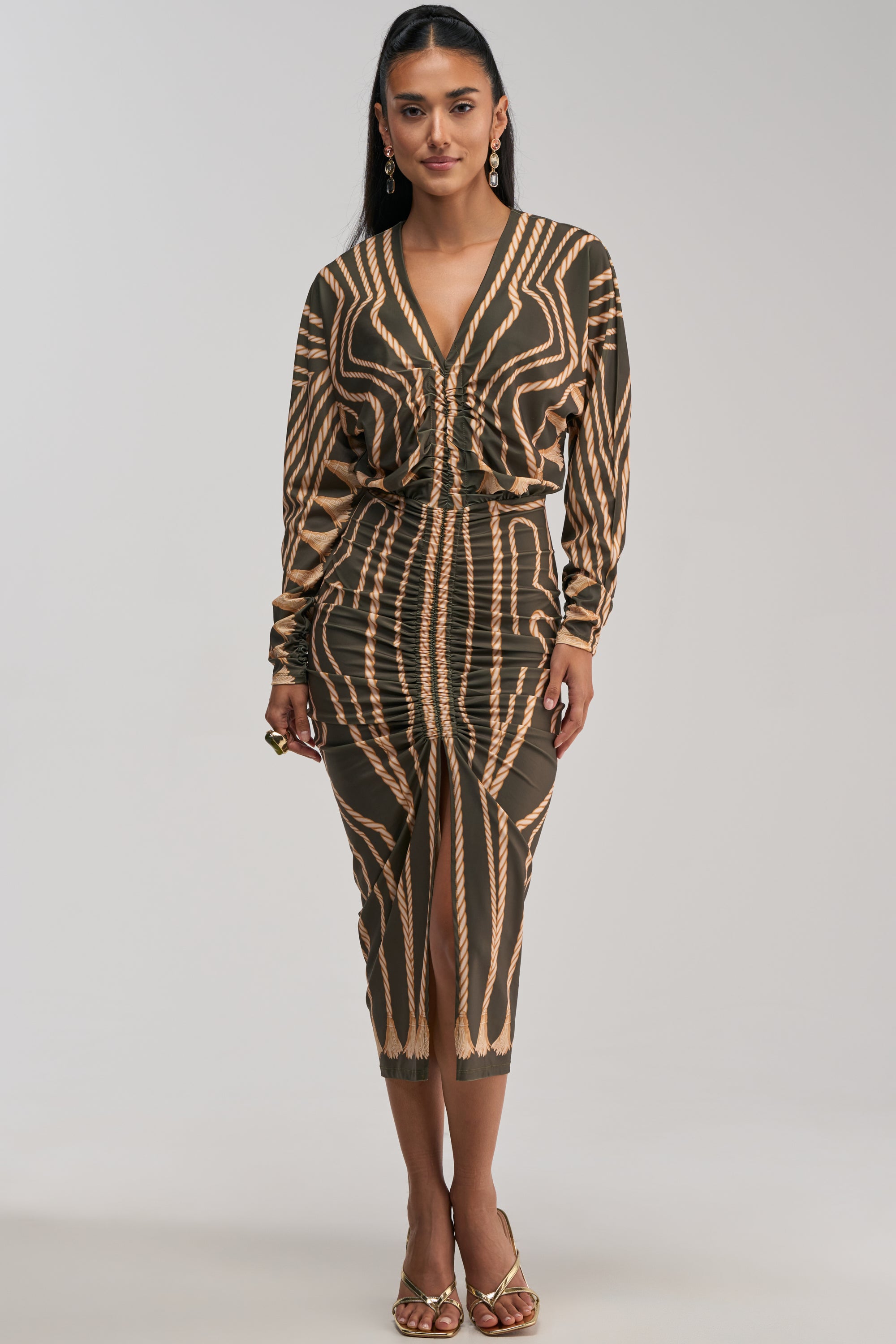A woman styled in the RESERVATIONS ONLY PRINTED MIDI DRESS IN OLIVE, featuring an olive green and gold abstract print, ruched detailing, V-neckline, long sleeves, and a front slit. She completes the look with gold heeled sandals and drop earrings.