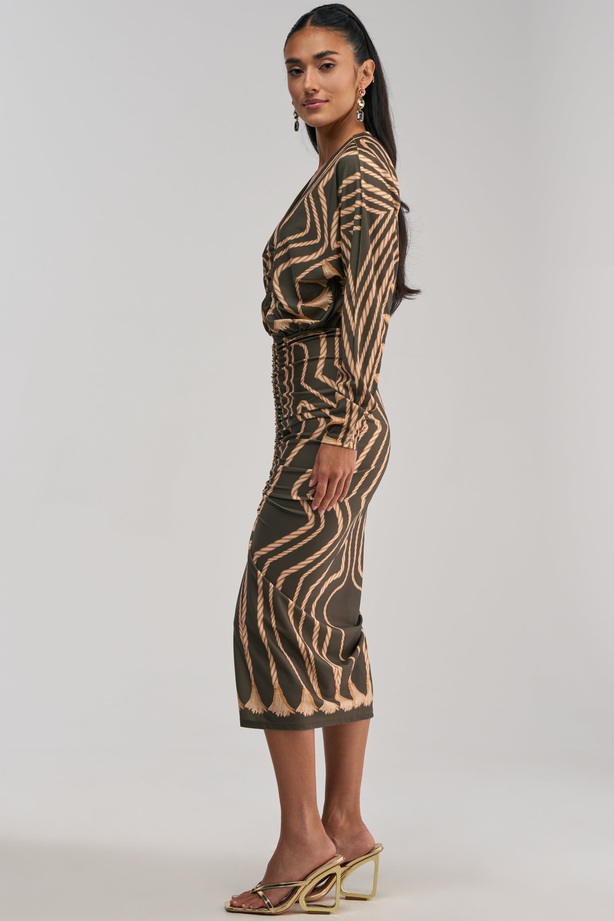 A woman stands sideways against a plain, light background wearing the RESERVATIONS ONLY PRINTED MIDI DRESS IN OLIVE, which features long sleeves, an abstract print in brown and beige tones with a fitted waist. She completes her look with gold hoop earrings and strappy gold heeled sandals.