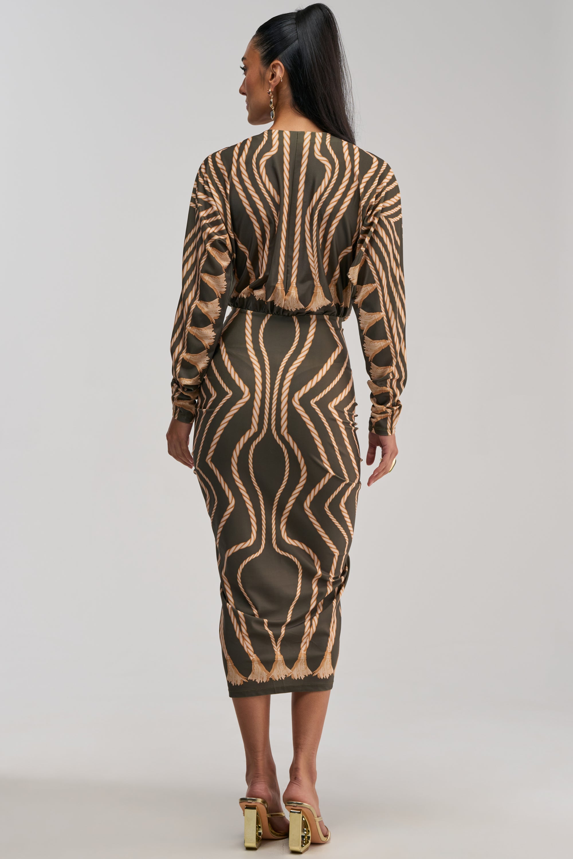 A woman stands facing away, wearing the RESERVATIONS ONLY PRINTED MIDI DRESS IN OLIVE, a fitted long-sleeve midi dress featuring bold tan and brown geometric patterns and ruched detailing. Her long dark hair is styled in a high ponytail, and she completes the look with gold heels against a plain, light-colored background.