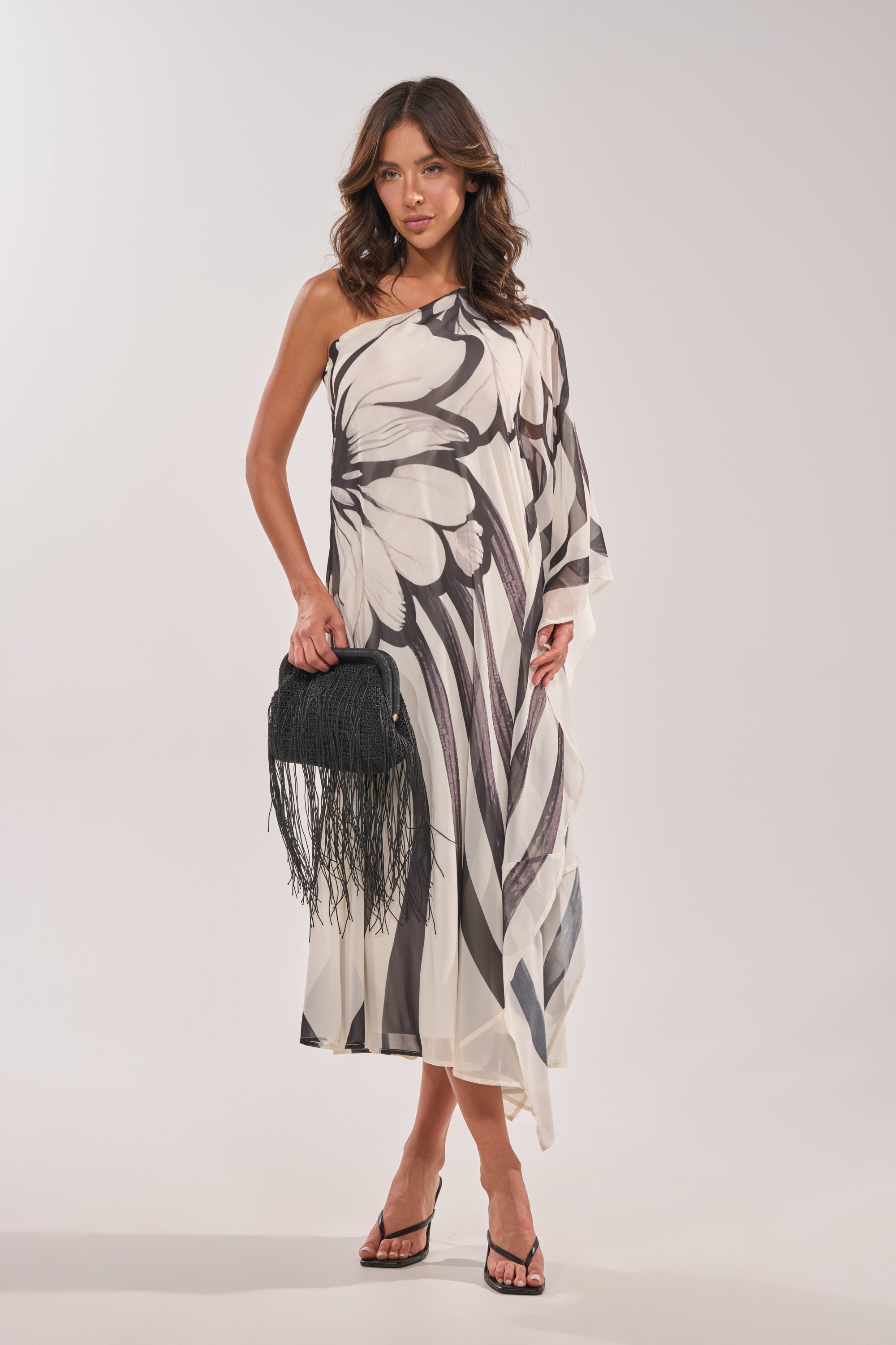 A woman models the TROPICAL NIGHTS ONE SHOULDER MAXI DRESS, featuring a white design with bold black butterfly wings. She stands against a plain background, accessorized with a black fringed handbag and black sandals. 1
