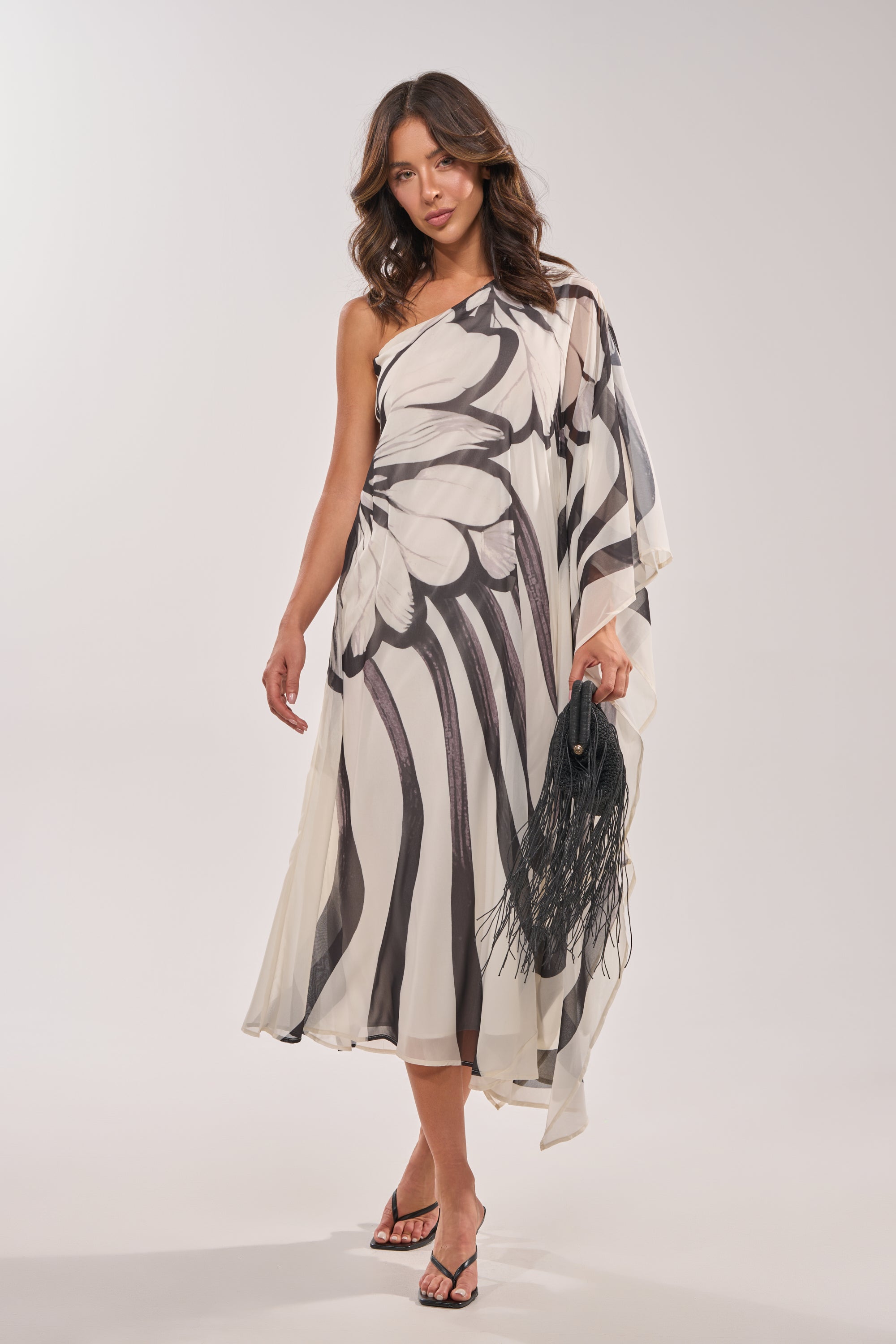 Wearing the TROPICAL NIGHTS ONE SHOULDER MAXI DRESS—a flowy black-and-white gown with an abstract wing-like pattern—a woman stands against a light background, accessorized with a fringed black clutch and black sandals. 2