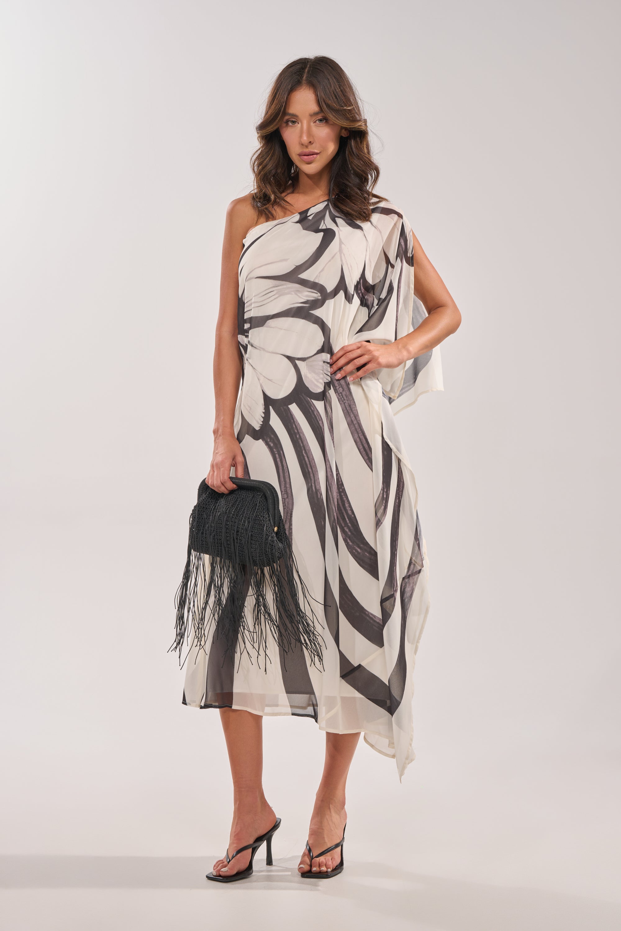 A woman poses against a plain background in the TROPICAL NIGHTS ONE SHOULDER MAXI DRESS featuring bold black abstract patterns. She accessorizes with a black fringed handbag, black heeled sandals, and wears her hair loose and wavy. 3