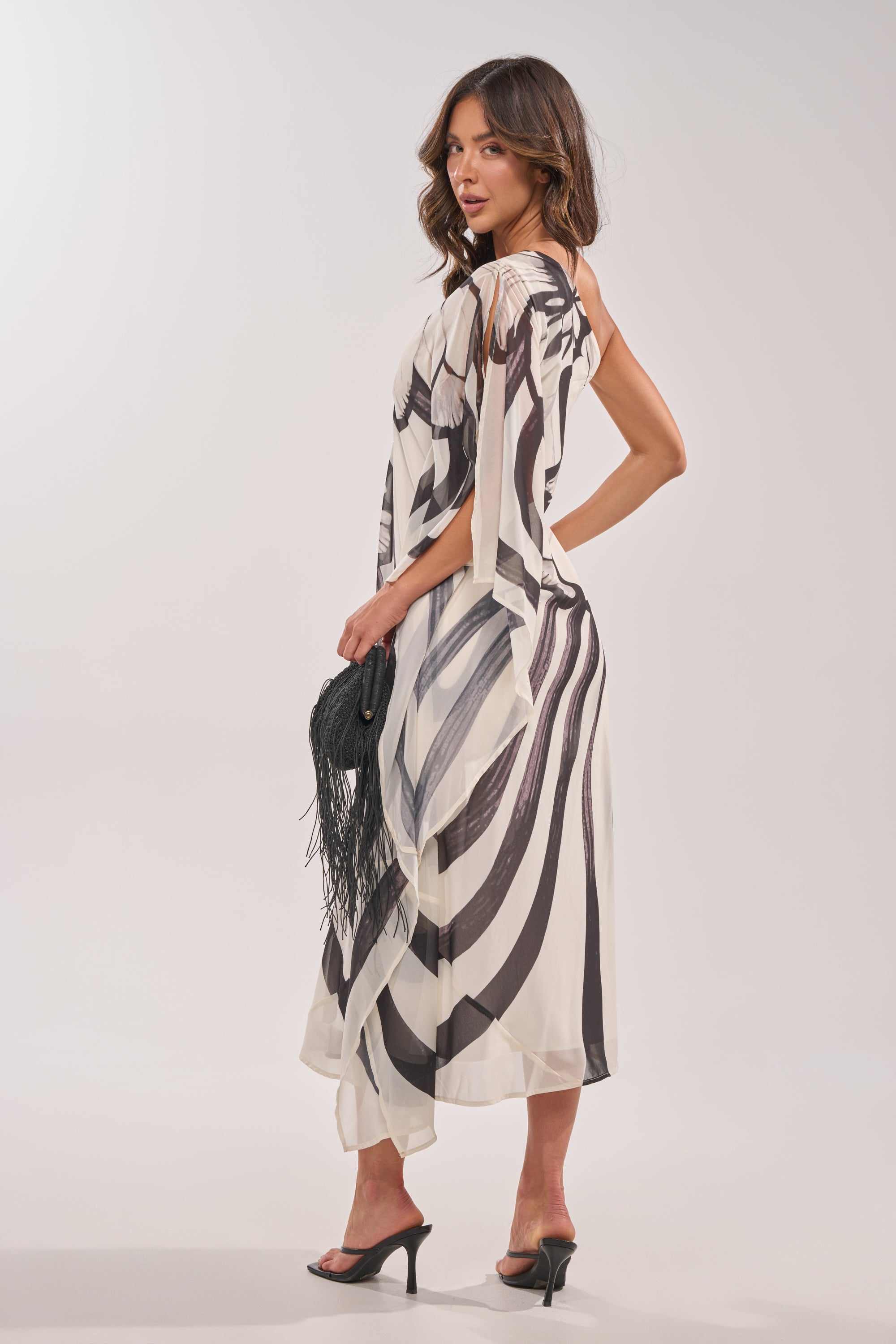 A woman in the TROPICAL NIGHTS ONE SHOULDER MAXI DRESS and black high-heeled sandals stands sideways, holding a black fringed handbag, against a plain white background. 4