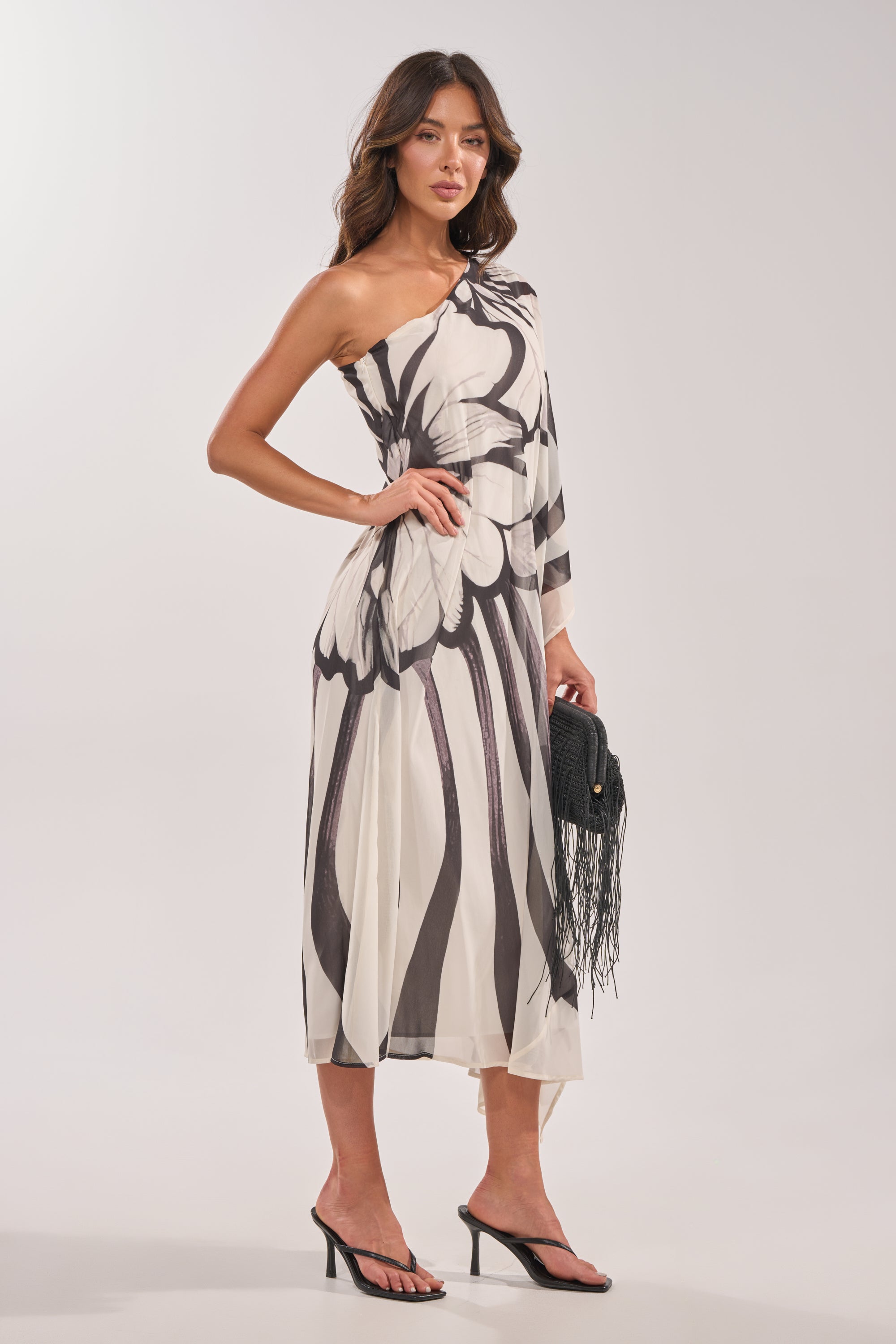 A woman wears the TROPICAL NIGHTS ONE SHOULDER MAXI DRESS with black heeled sandals and a black fringed clutch, standing against a plain light background. 5