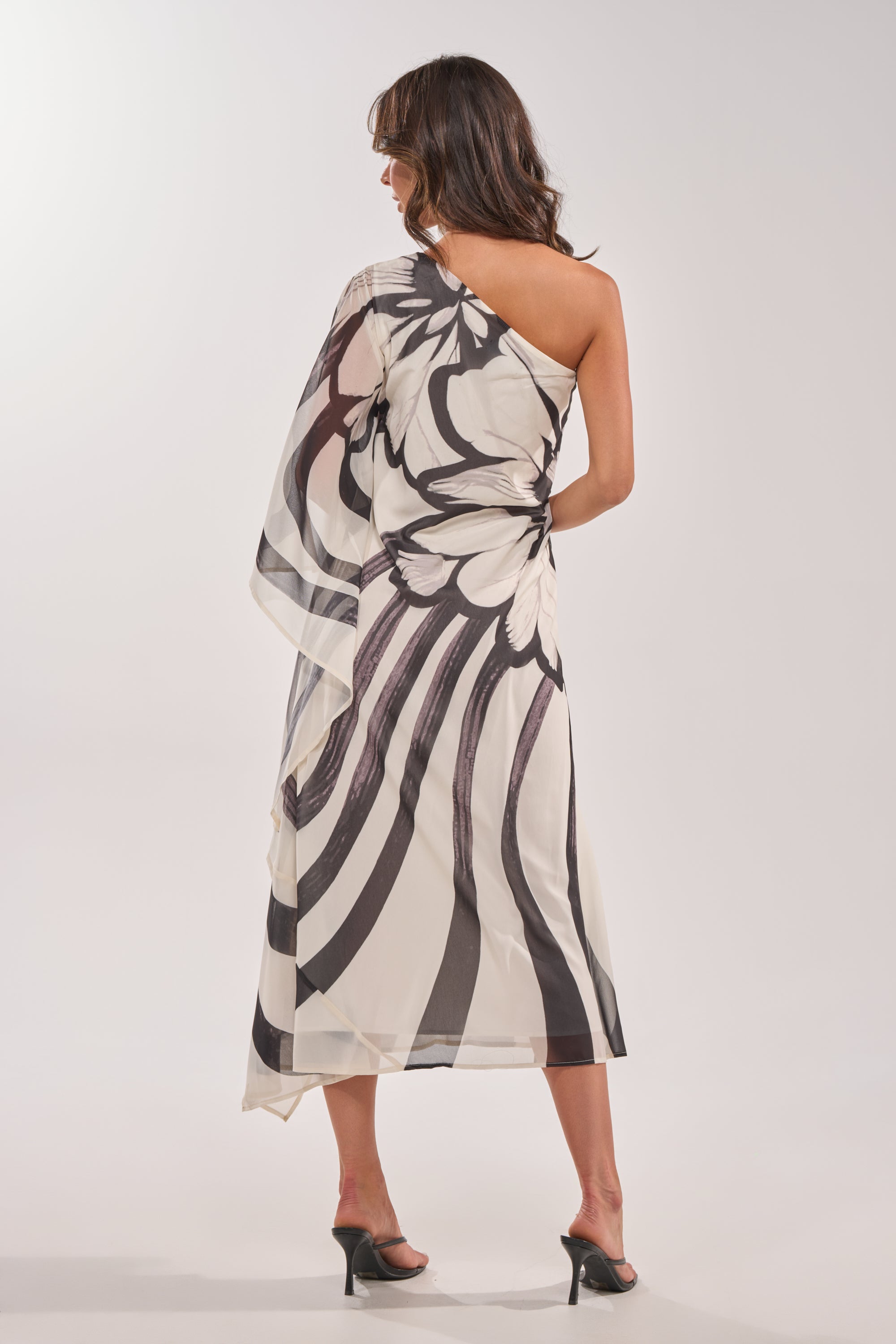 A woman stands facing away, wearing the TROPICAL NIGHTS ONE SHOULDER MAXI DRESS with a bold black and white botanical print, paired with black heeled sandals against a plain light background. 6