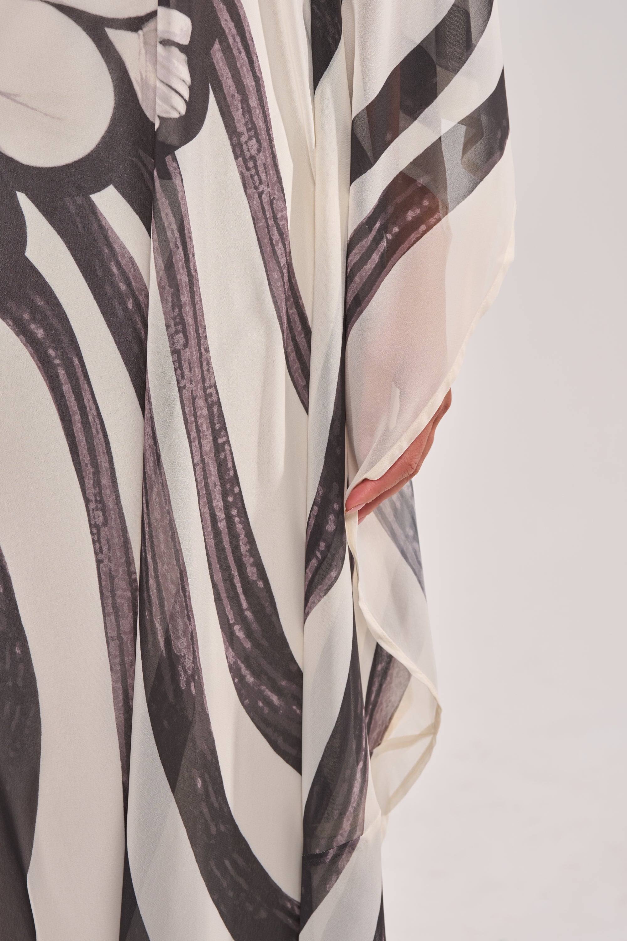 A close-up of someone in the TROPICAL NIGHTS ONE SHOULDER MAXI DRESS, featuring flowing black-and-white abstract patterns and a sheer sleeve, with their hand partially visible against a plain light background. 7