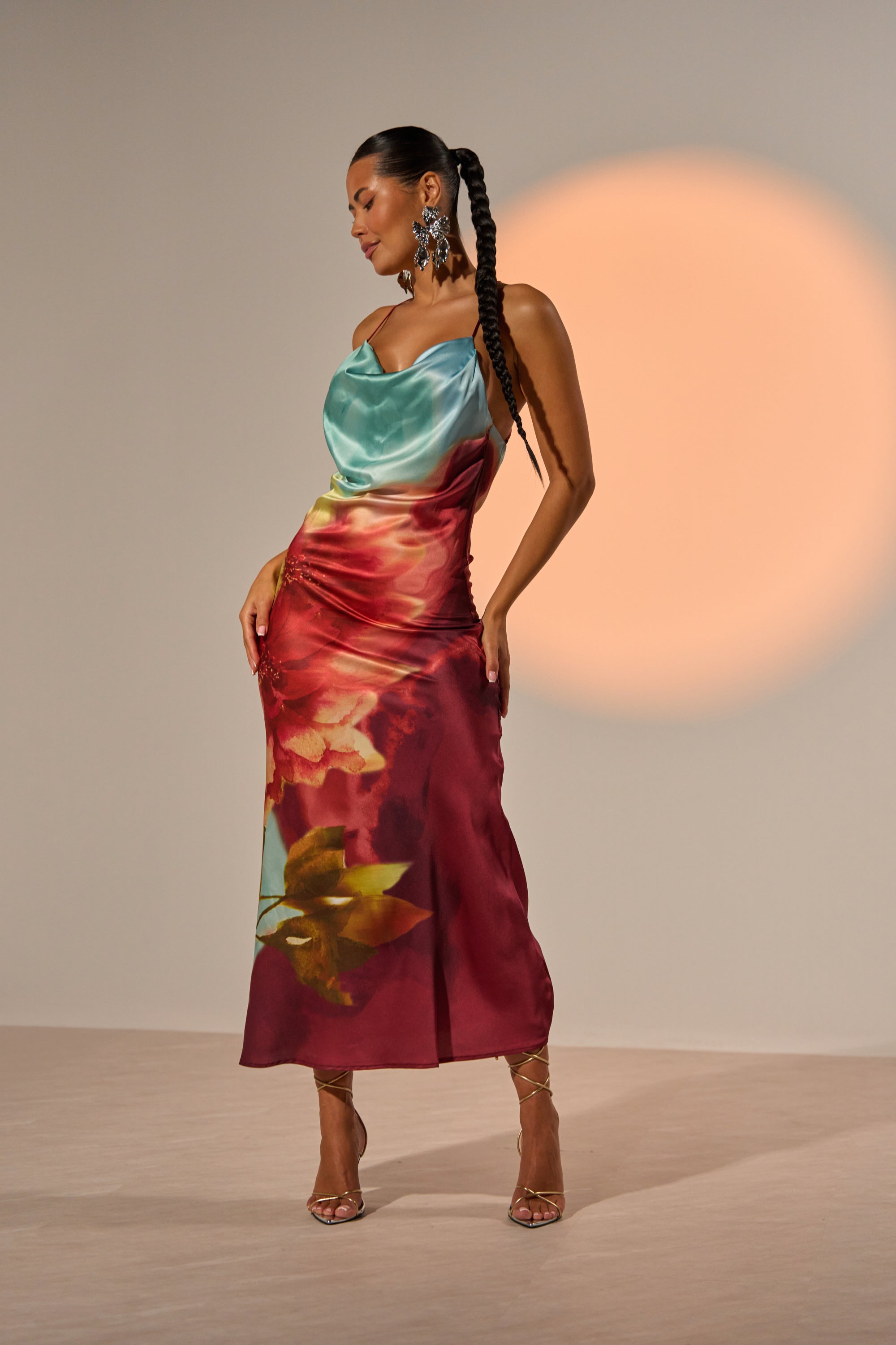 A woman showcases the WORK OF ART SATIN MAXI DRESS, featuring a teal cowl neckline and a red-and-yellow floral skirt, styled with statement earrings, gold heels, and a braided ponytail before an orange circular spotlight. 1