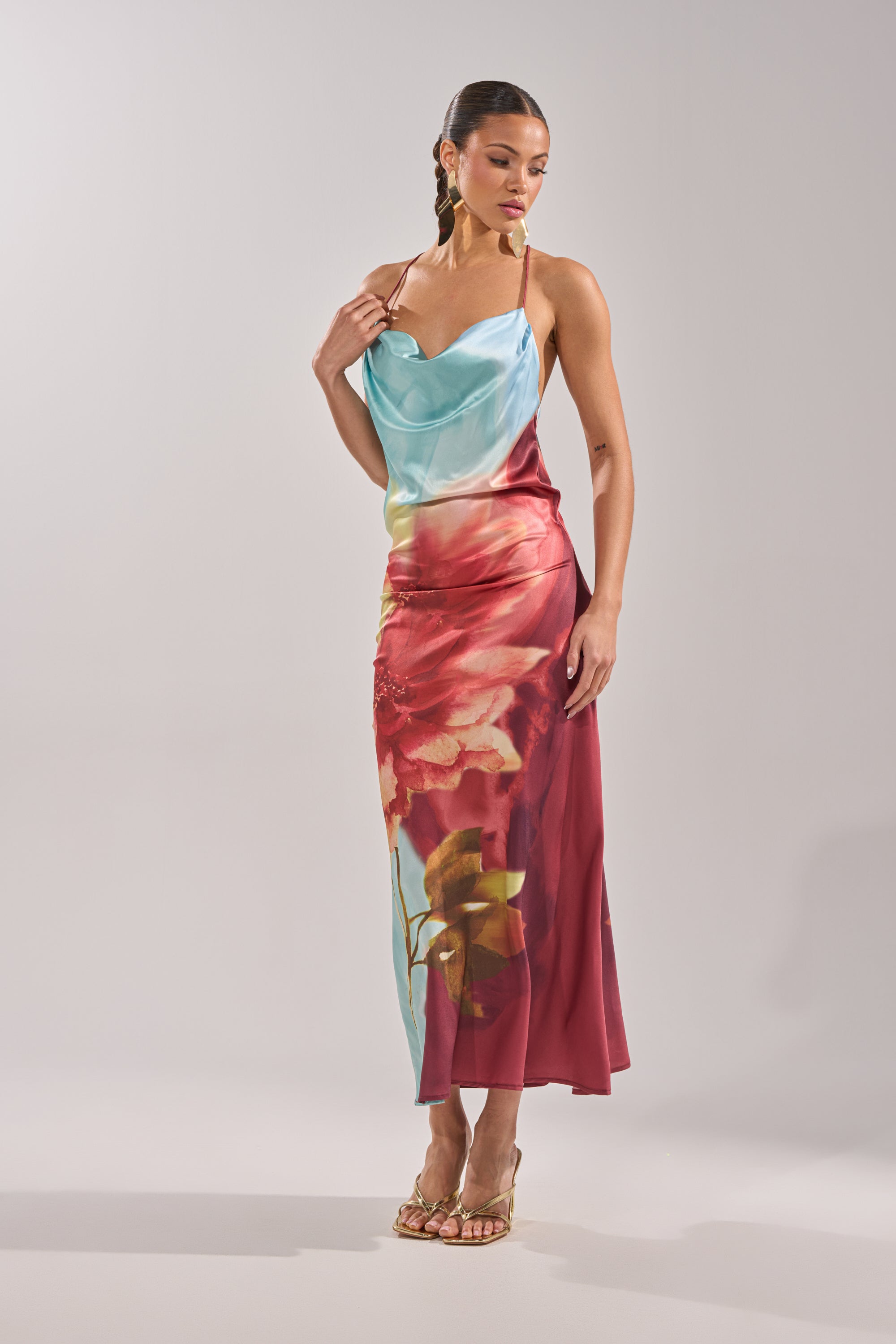 A woman wears the WORK OF ART SATIN MAXI DRESS, a sleeveless maxi with a cowl neckline, featuring a light blue gradient top and large red and yellow floral patterns on the ankle-length satin skirt. 4