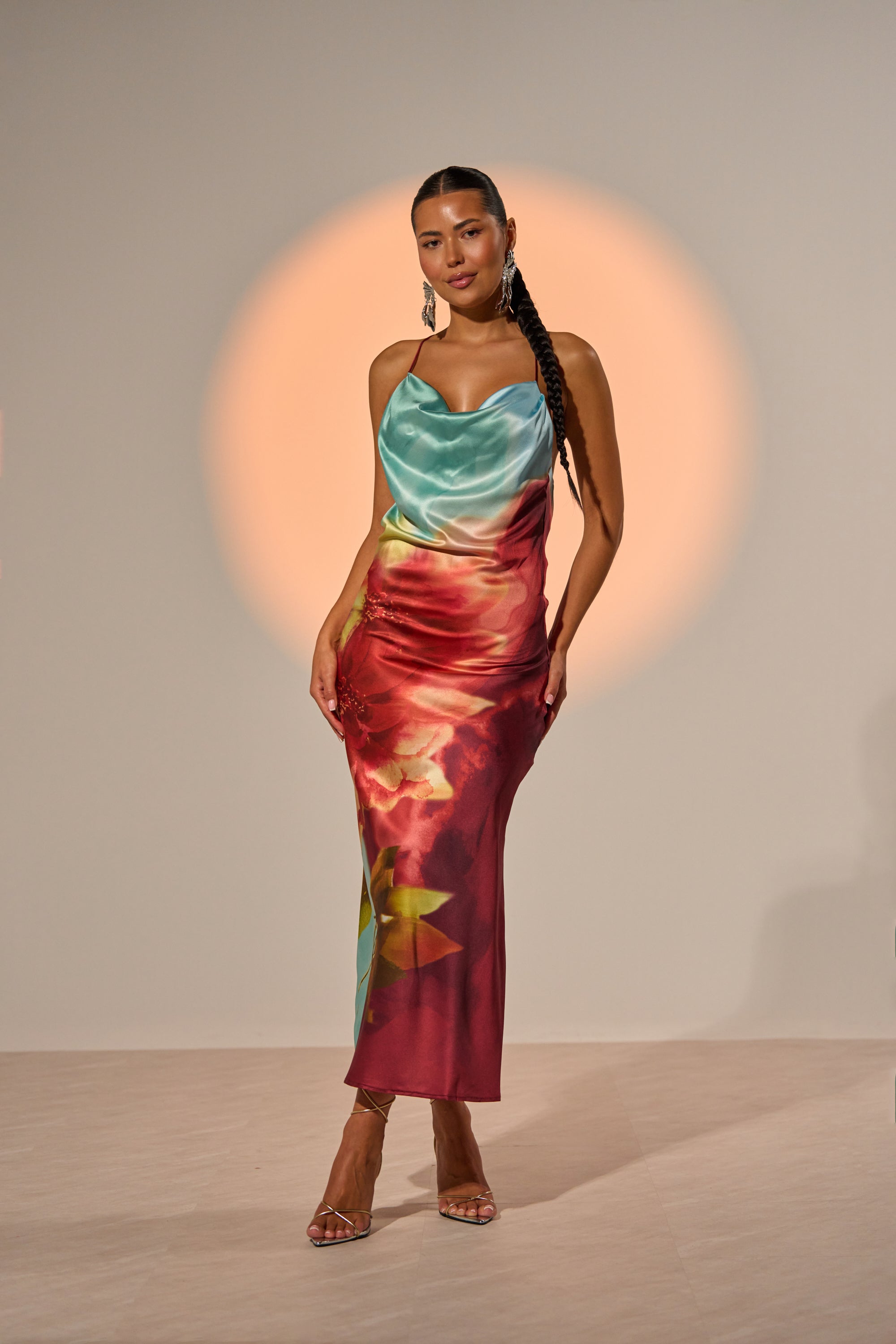 A woman with long braids confidently poses in the WORK OF ART SATIN MAXI DRESS, showcasing a vibrant blue top and red floral skirt, set against a plain backdrop illuminated by a soft, circular orange light. 2