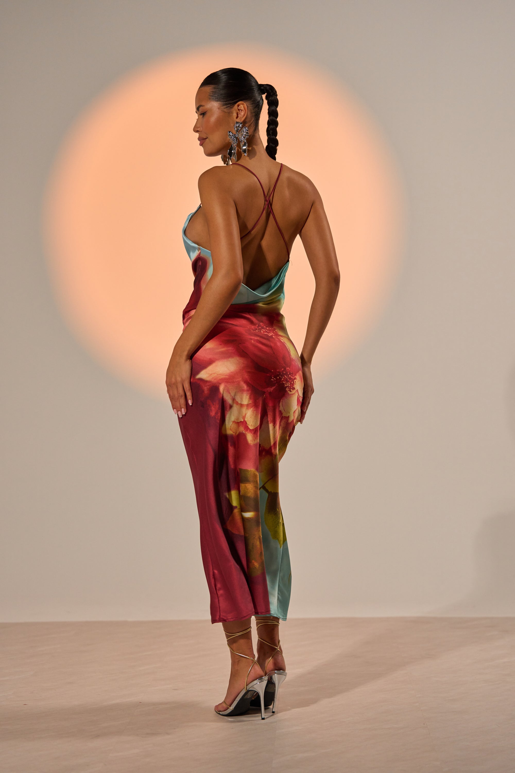 A woman with a long braid showcases the WORK OF ART SATIN MAXI DRESS, a colorful maxi with bold floral print on the back. She poses in profile under an orange spotlight, accessorized with strappy heels and statement earrings. 3