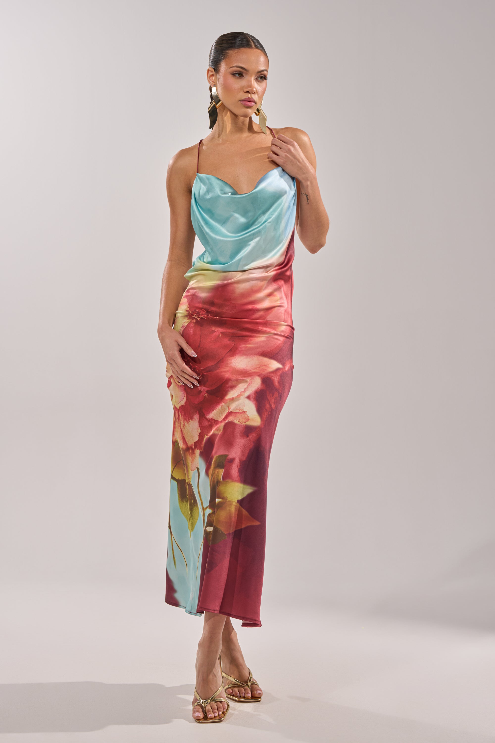 A woman poses in the WORK OF ART SATIN MAXI DRESS featuring a floral print, draped turquoise bodice, and red skirt. She completes her elegant look with gold heels, statement earrings, and slicked-back hair. 6