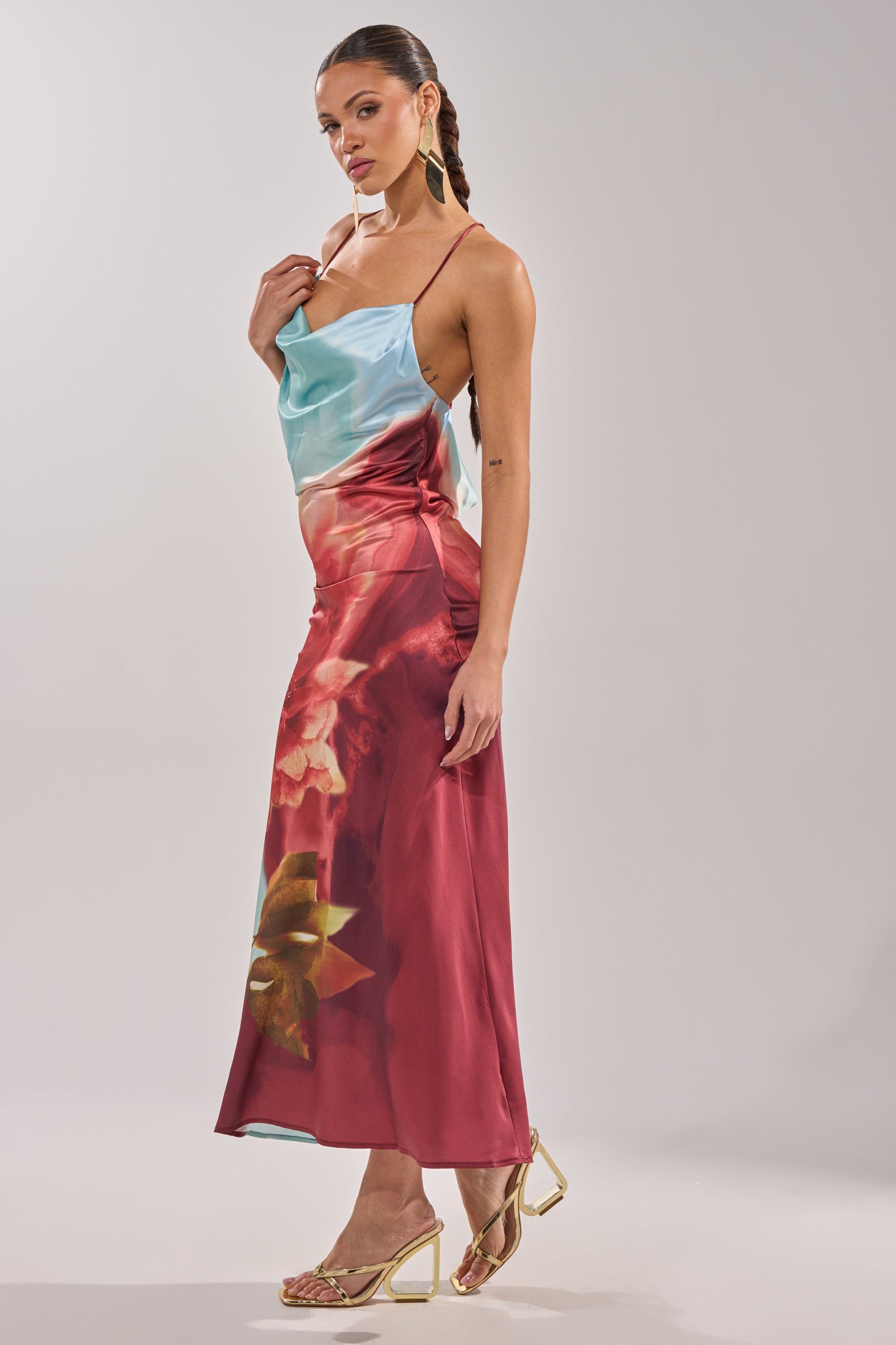 A woman stands sideways wearing the WORK OF ART SATIN MAXI DRESS with blue and red floral patterns, paired with gold hoop earrings, gold heeled sandals, and a braided ponytail. 7
