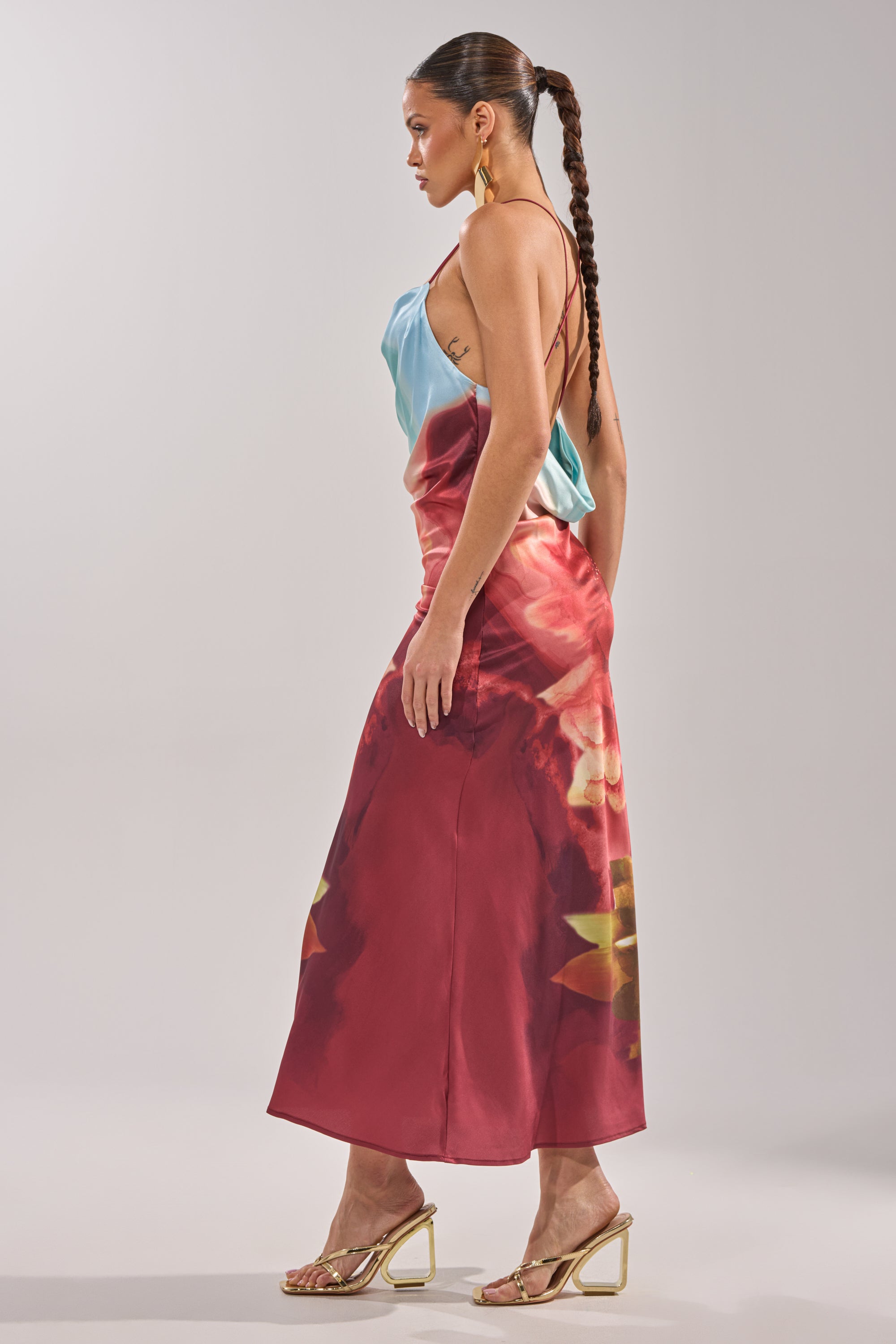 A woman with long braided hair and gold heels wears the WORK OF ART SATIN MAXI DRESS—a blue and red floral, spaghetti strap maxi—standing in profile against a plain light background. 8