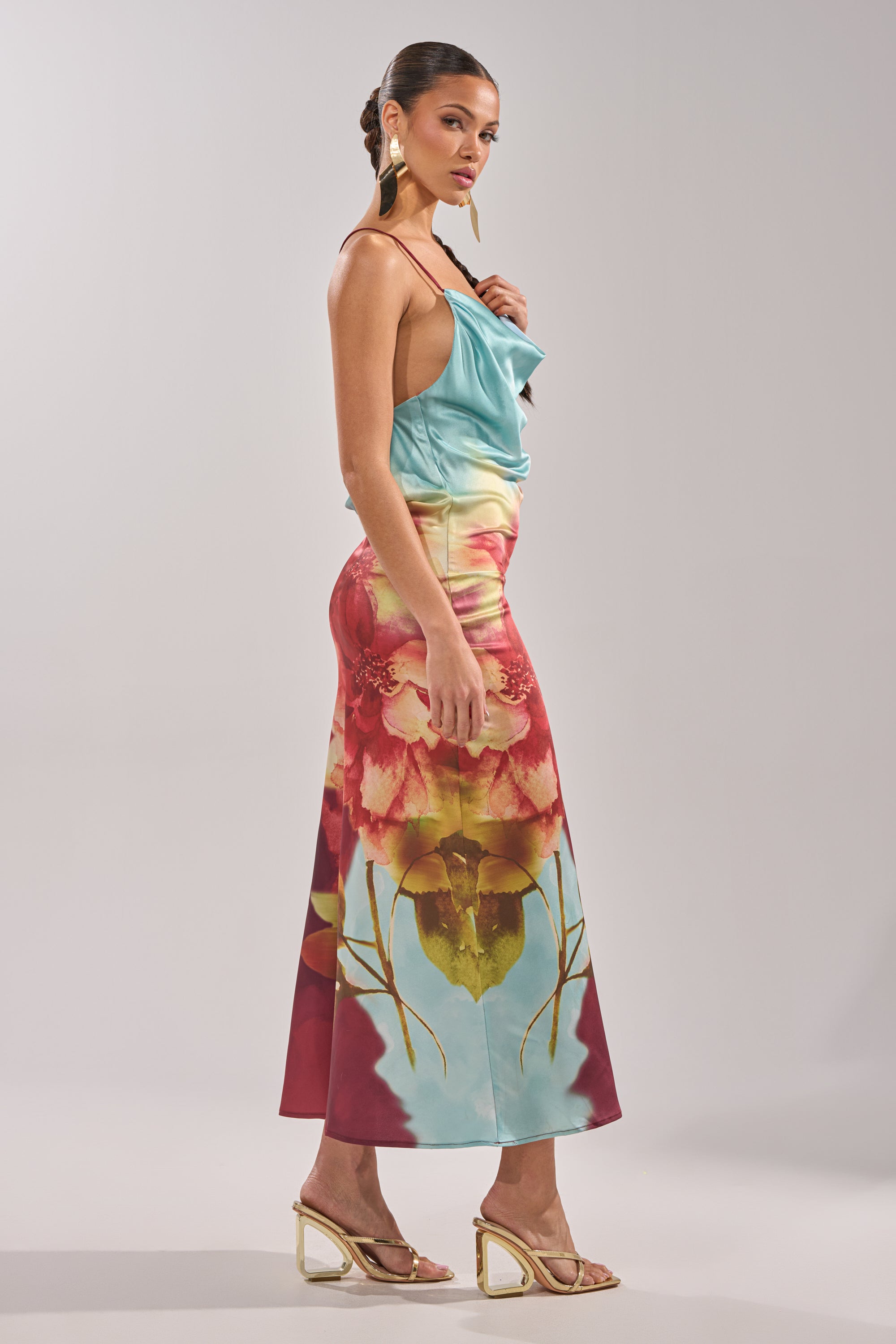 A woman stands in profile against a plain background, wearing the WORK OF ART SATIN MAXI DRESS with gold earrings and strappy heels. Her hair is styled in a sleek updo. 9