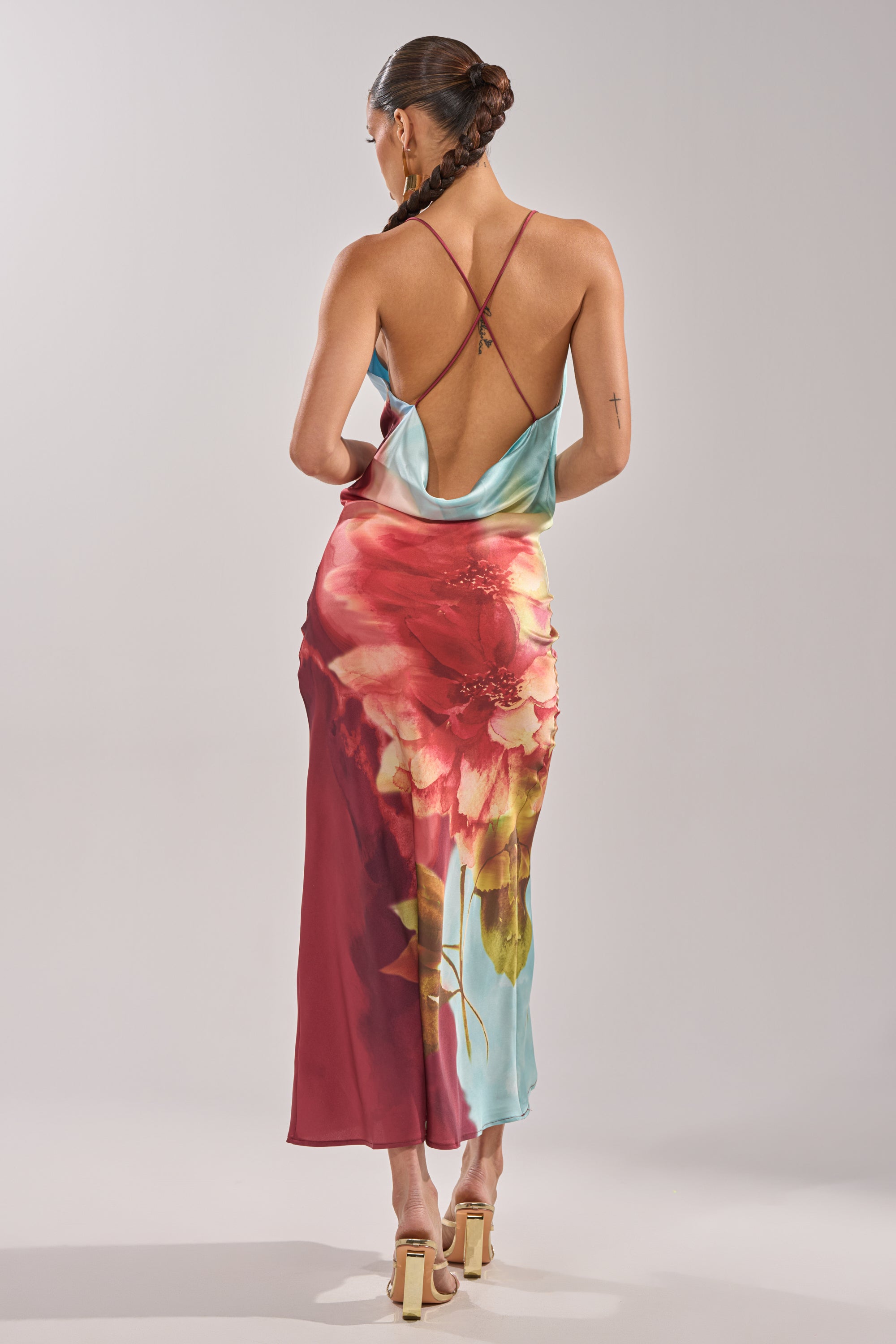 A woman with braided hair models the WORK OF ART SATIN MAXI DRESS—a backless, strappy maxi with a bold red and aqua floral print—paired with heeled sandals against a plain light background. 10