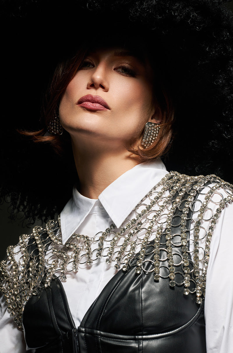 A woman in a large black hat and statement earrings poses confidently, with her hair styled in soft waves, dressed in a white shirt embellished with the RICH BISH STATEMENT NECKLACE and a leather garment.