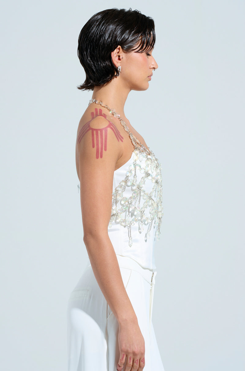 A person with short black hair stands in profile, showcasing a striking festival look. They are wearing the FANTASY DROP RAZOR CRYSTAL BODY CHAIN TOP, adorned with intricate beaded detailing. A bold, artistic red design resembling an eye with drips is painted on their shoulder, echoing the edgy Y2K style.