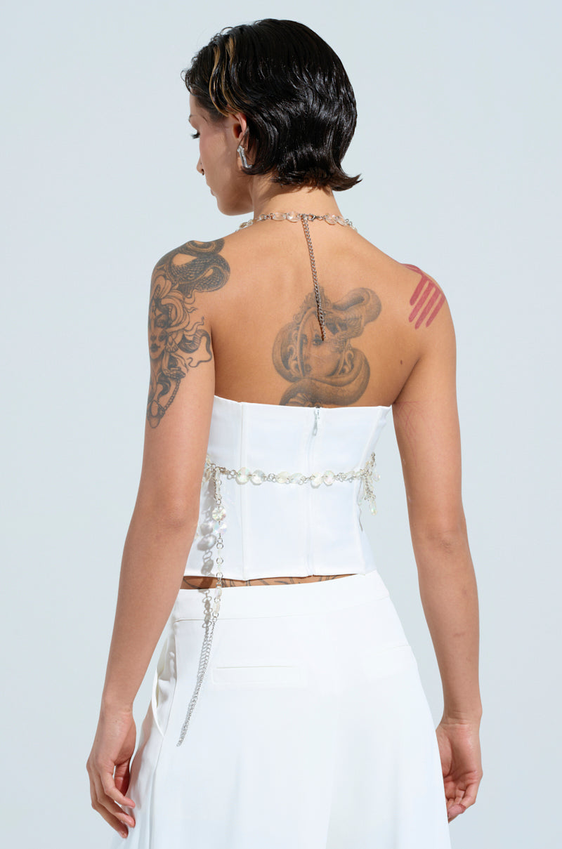 A person with short dark hair facing away is wearing a FANTASY DROP RAZOR CRYSTAL BODY CHAIN TOP and white pants. Tattoos adorn their shoulder and back, enhancing the Y2K style flair against the light gray background.