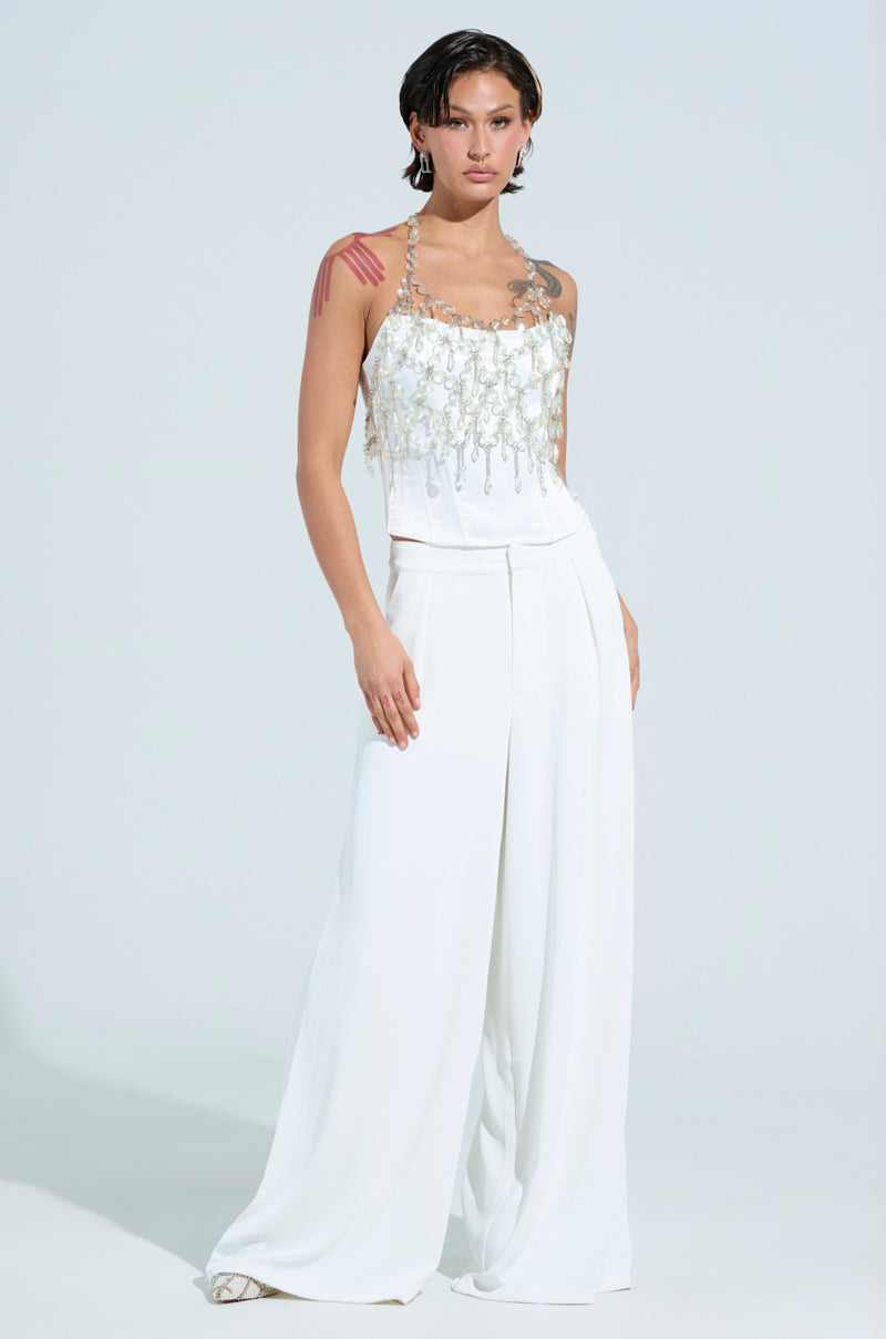 Against a plain background, a person confidently poses in the FANTASY DROP RAZOR CRYSTAL BODY CHAIN TOP, part of a stylish white outfit. The top showcases intricate beadwork, complemented by wide-legged pants that add flair to the look. Channeling a Y2K aesthetic with short, slicked-back hair, they strike a pose with one hand on their hip.