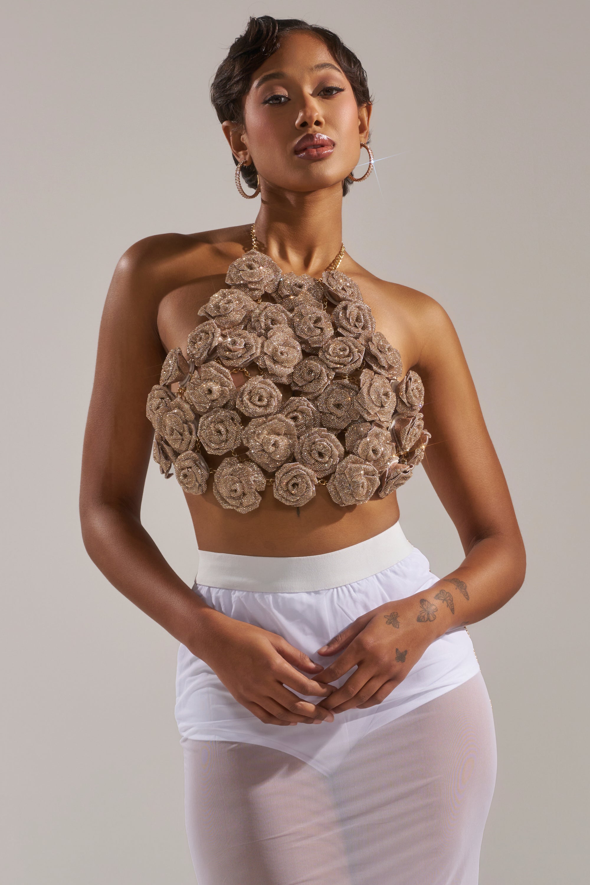 A woman poses confidently in the ROSES BRALETTE, featuring a halter neckline and textured, rose-shaped embellishments, paired with a sheer white skirt against a plain gray background. 0