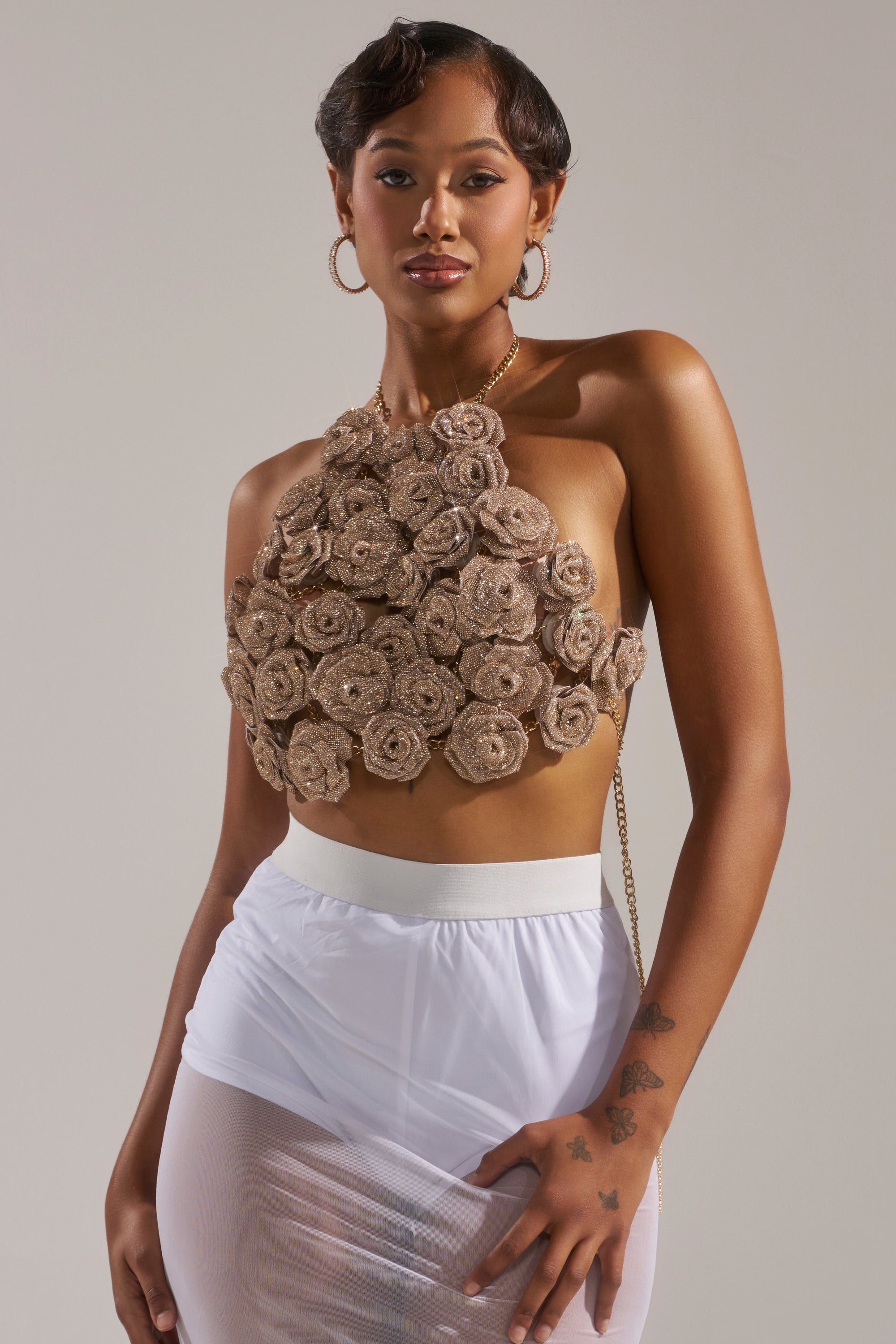 A woman poses confidently in the ROSES BRALETTE, a beige halter crop top with rose-shaped embellishments, paired with a sheer white skirt. She accessorizes with hoop earrings, short hair, floral forearm tattoos, and stands against a neutral background. 2